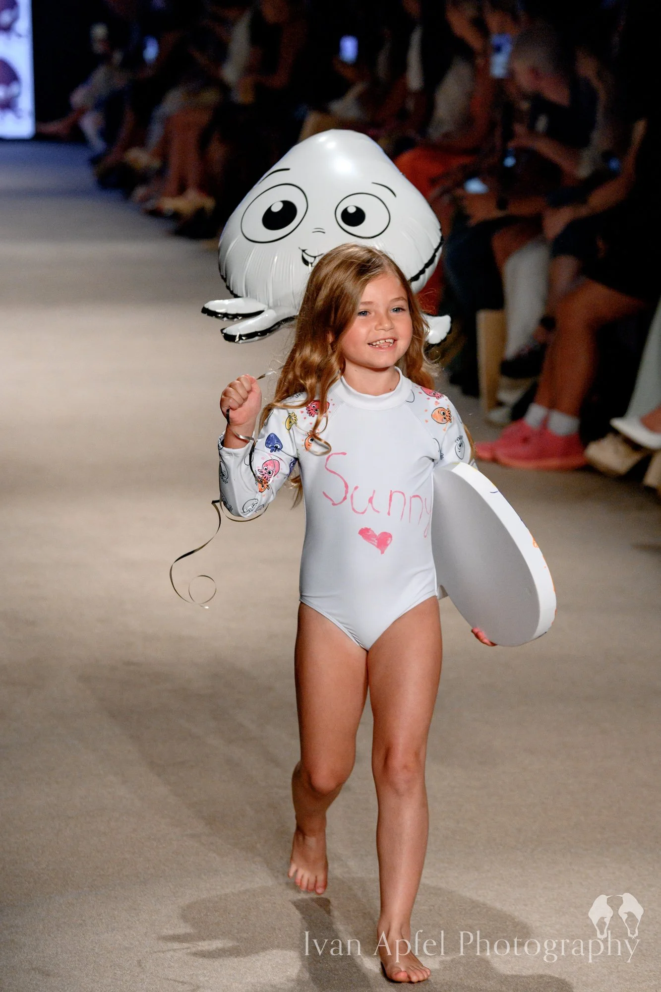 Young girl walking barefoot on a runway, wearing a white swimsuit with colorful character prints, holding a white surfboard, and carrying a large helium balloon with a cartoon face and big eyes behind her, during a fashion show.