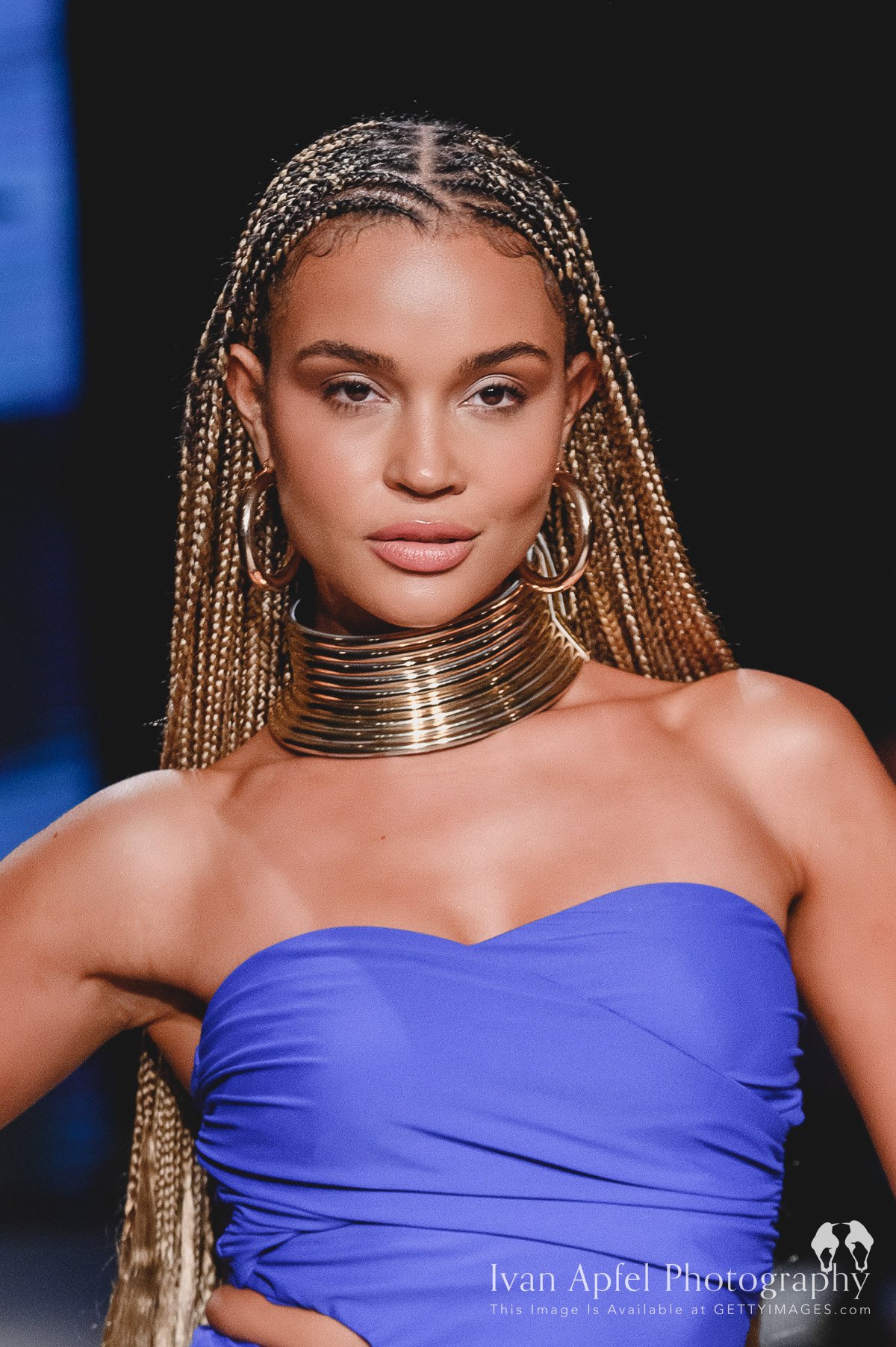 Close-up of a woman with braided hair wearing large gold hoop earrings, layered gold choker, and a strapless blue dress, against a dark background.