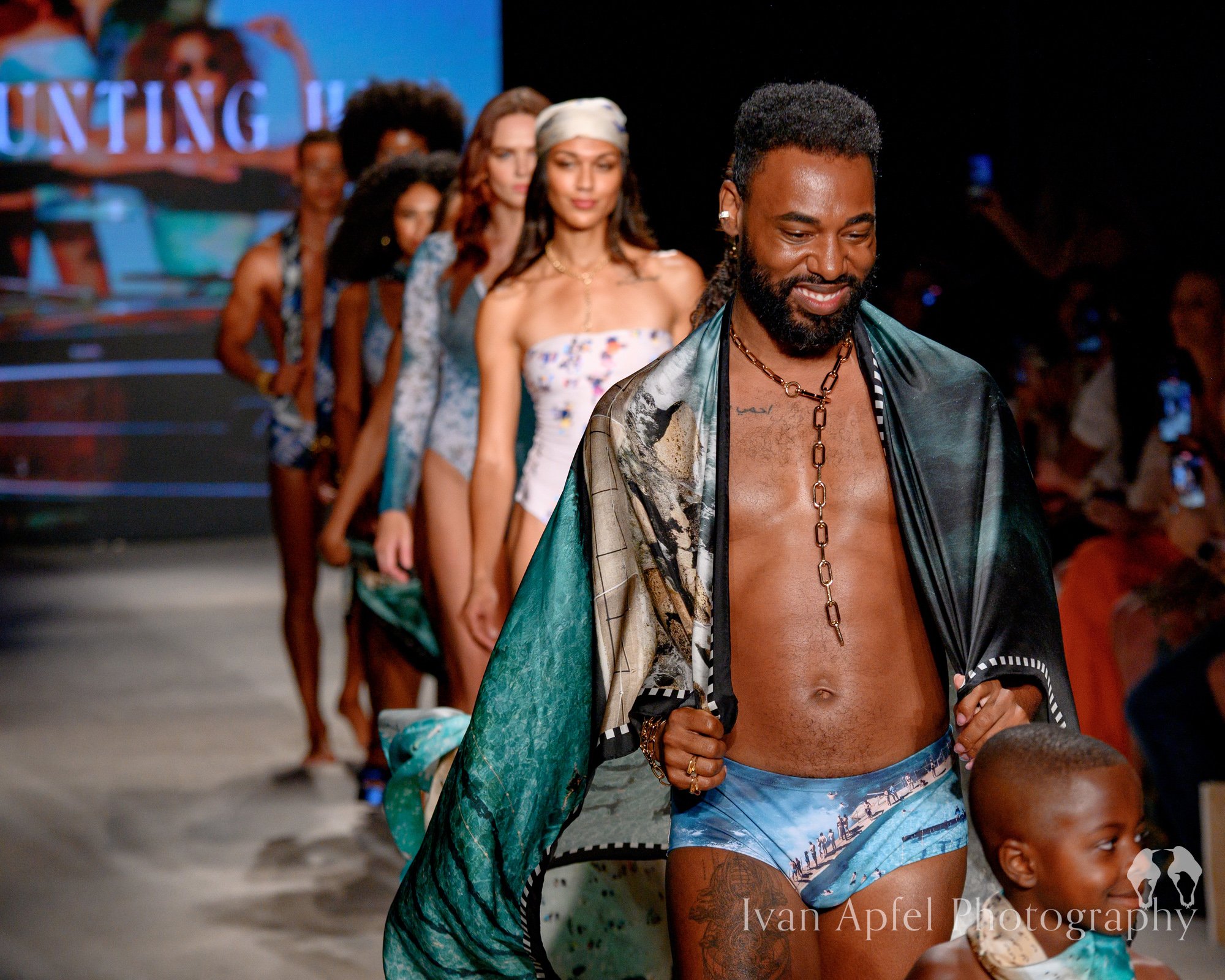 Man walking on runway at fashion show wearing a silky, patterned cape, blue swim trunks, and a chain necklace, followed by female models in swimsuit outfits.