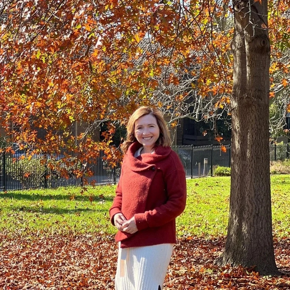 A woman in a red coat standing outdoors on autumn leaves in front of an orange and red tree.