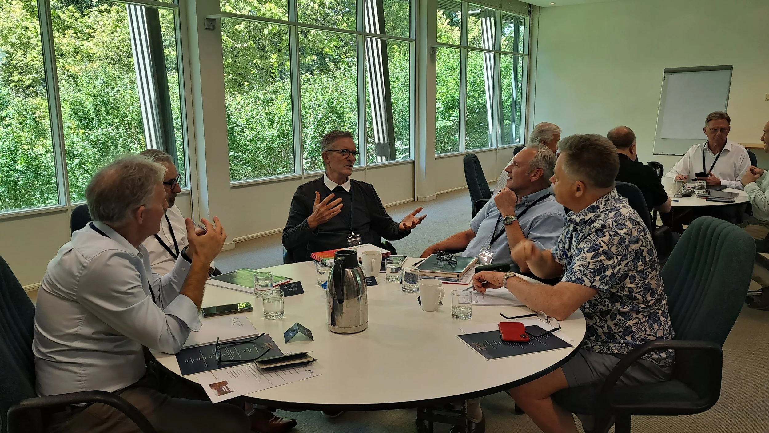 Group of professionals having a discussion around a conference table in a bright room with large windows and a view of green trees outside.