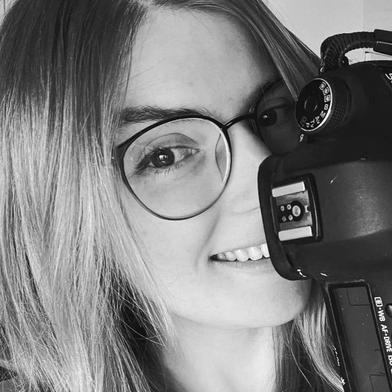 A young woman with glasses smiling while taking a selfie with a DSLR camera.