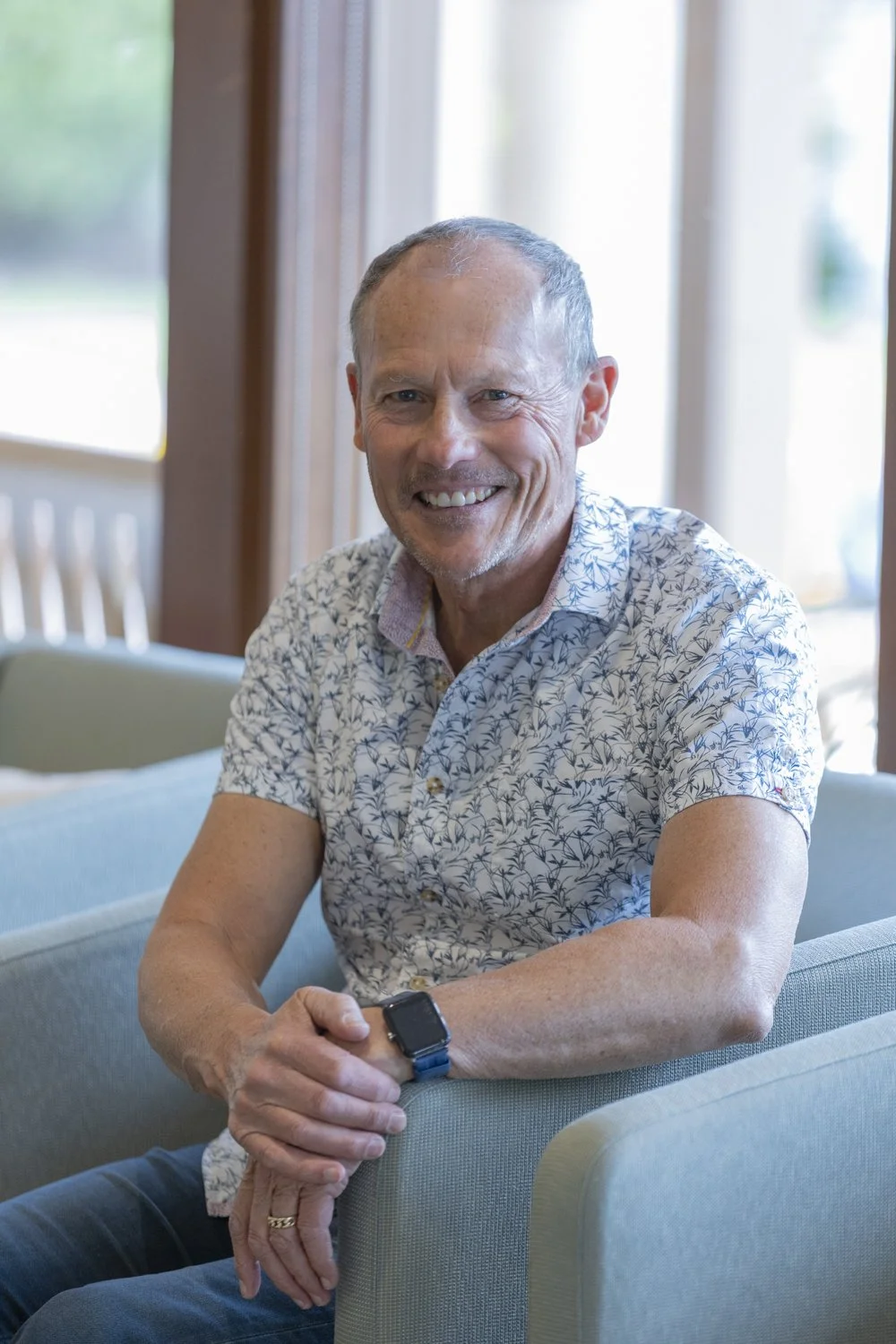 Smiling middle-aged man with short gray hair sitting on a light blue armchair indoors, wearing a short-sleeved floral shirt and a smartwatch, with a large window and green outdoor scenery in the background.