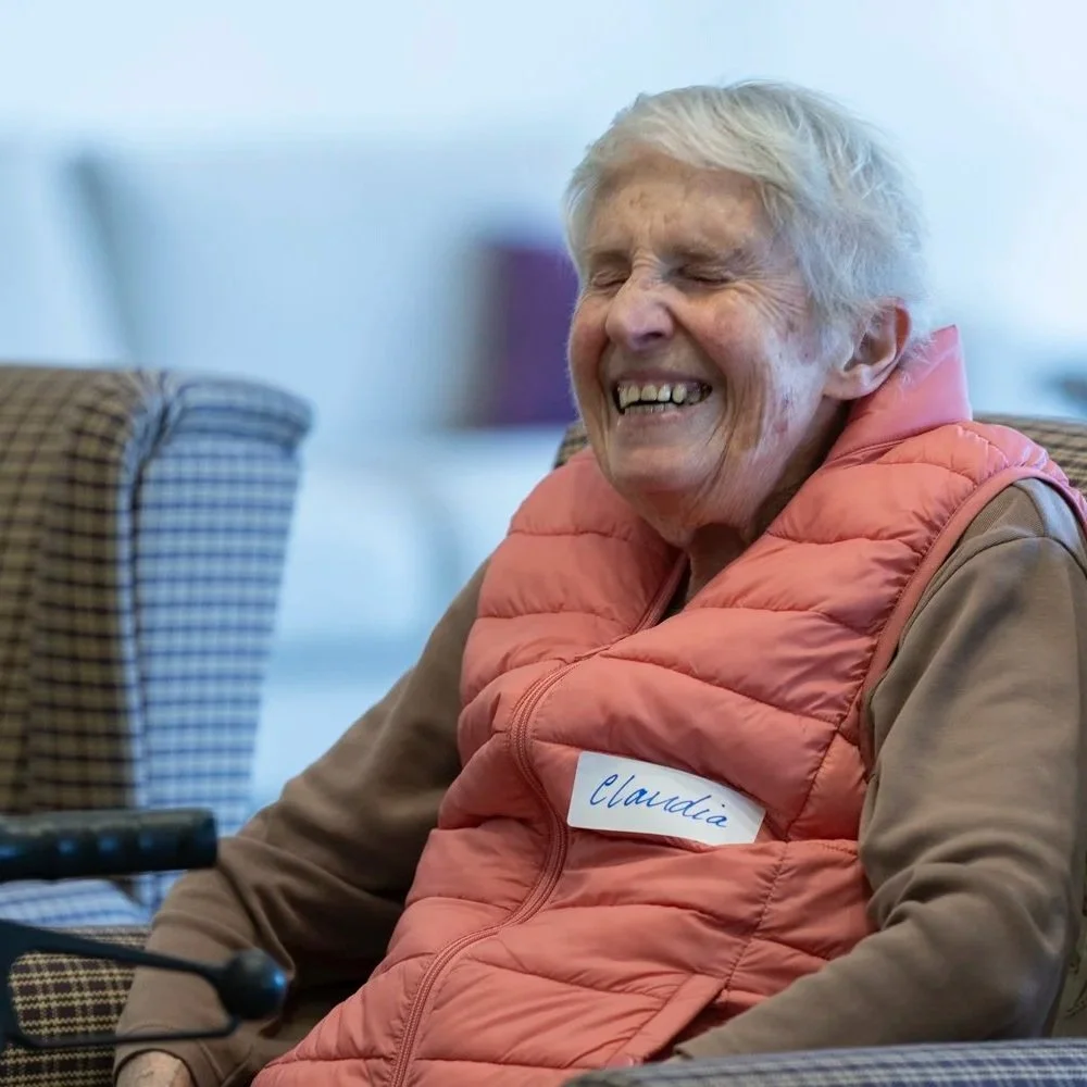 An elderly woman with short white hair, smiling and laughing, wearing a tan shirt and an orange vest with a name tag that says 'Claudia,' sitting in a comfortable chair.