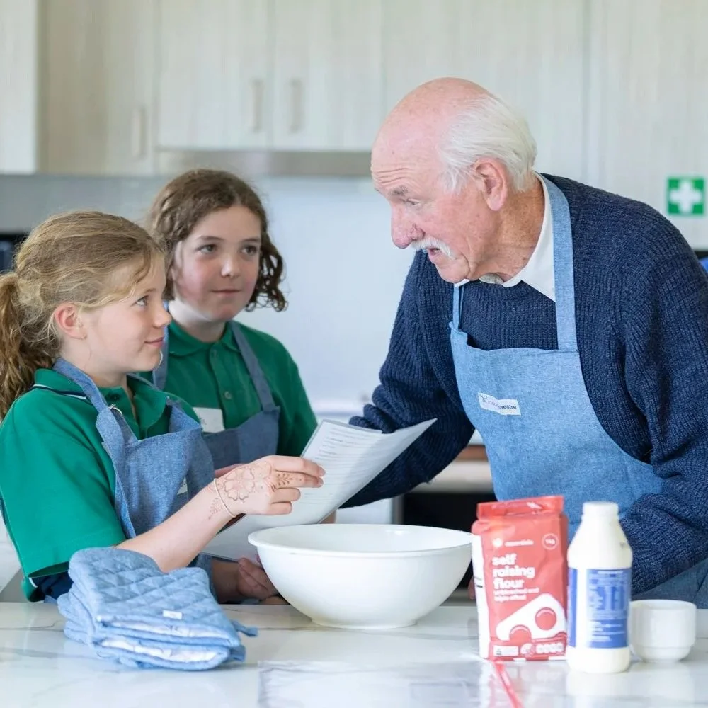 An elderly man and two young girls wearing aprons are in a kitchen. The man is showing a recipe on a sheet of paper to the girls, who are attentively listening. There are baking ingredients, including flour and milk, on the counter.