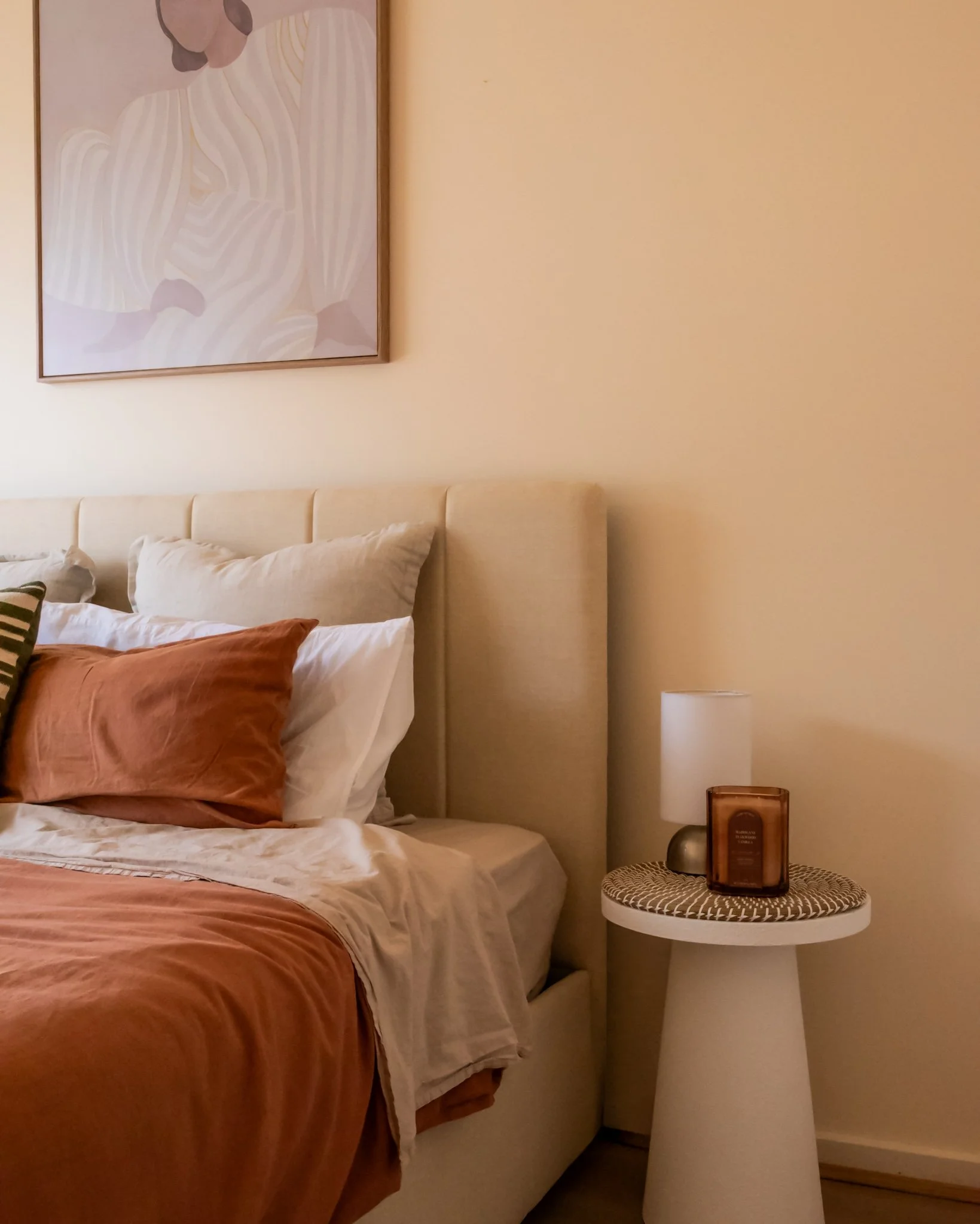 A cozy bedroom corner with a beige upholstered headboard, layered pillows, a textured orange blanket, a side table with a lamp and decorative items, and a framed abstract artwork on the wall.