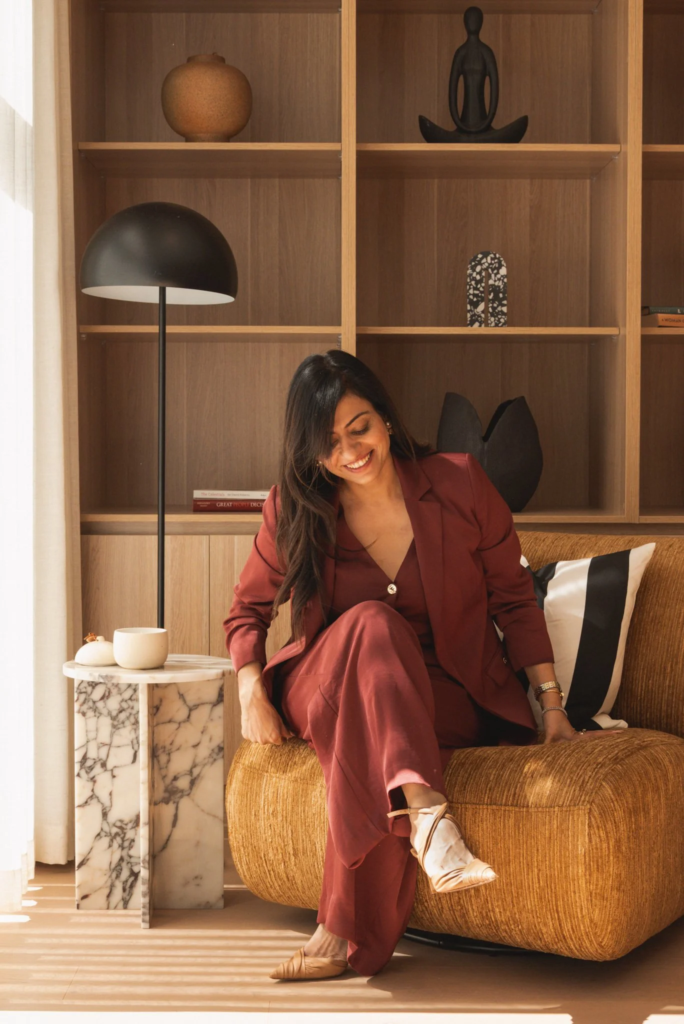 A woman in a maroon suit sitting on a mustard-colored sofa, smiling, in a modern living room with a wooden bookshelf and a marble side table.