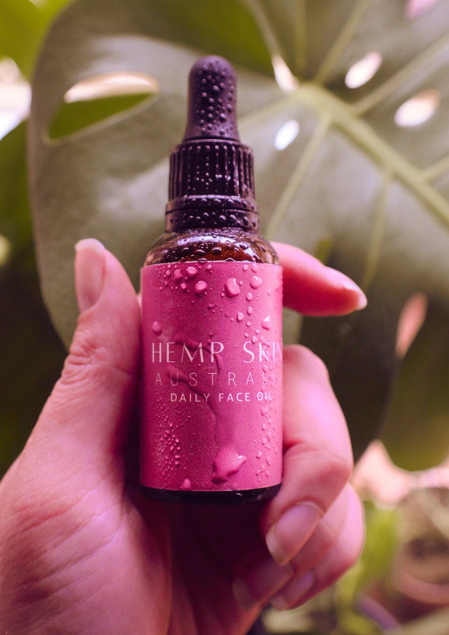 A hand holding a small glass bottle of hemp skin daily face oil with droplets of water on it, against a leafy background.