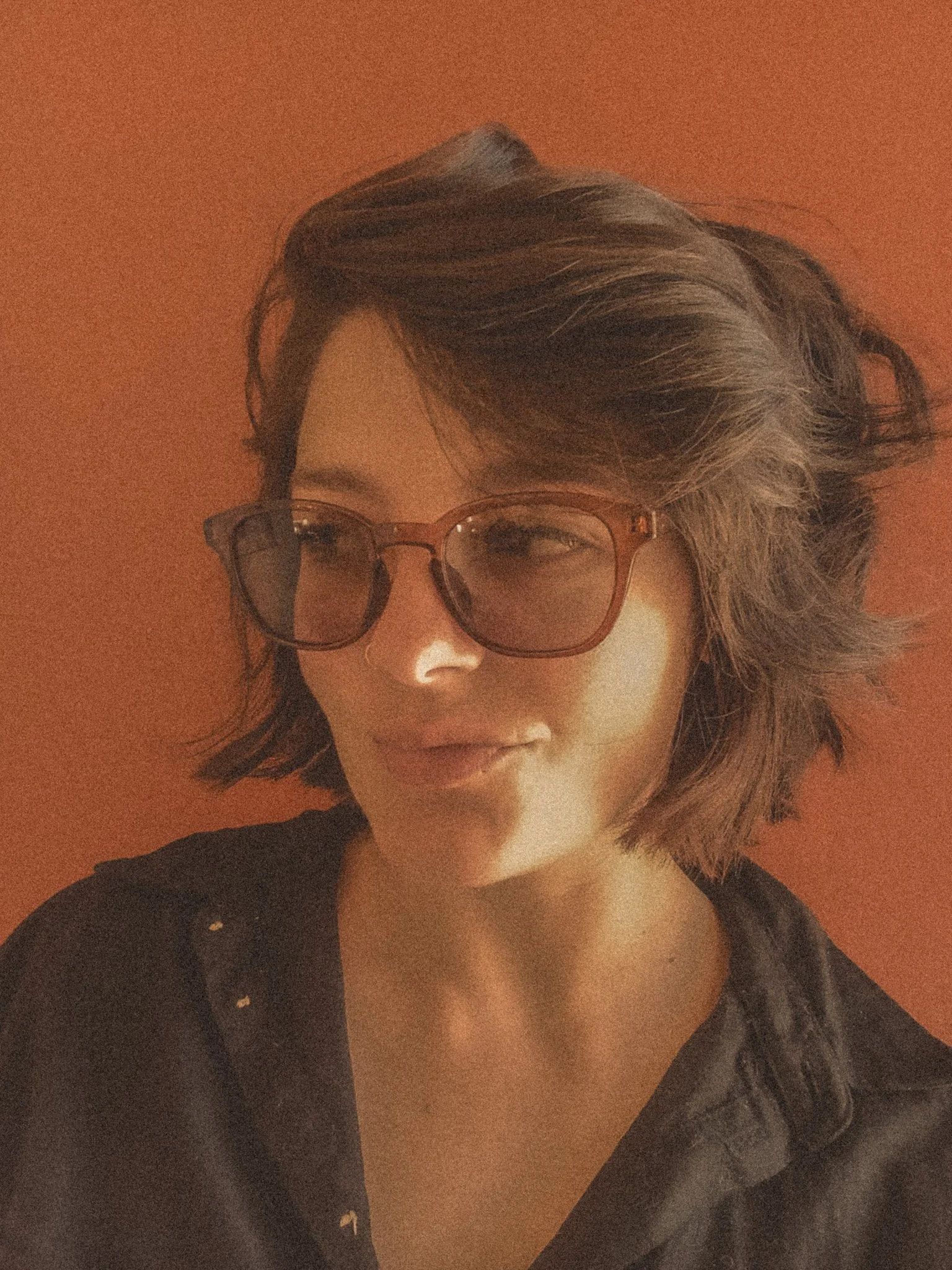 A woman with short, wavy hair wearing large, brown glasses and a black shirt, standing against a reddish-orange background.