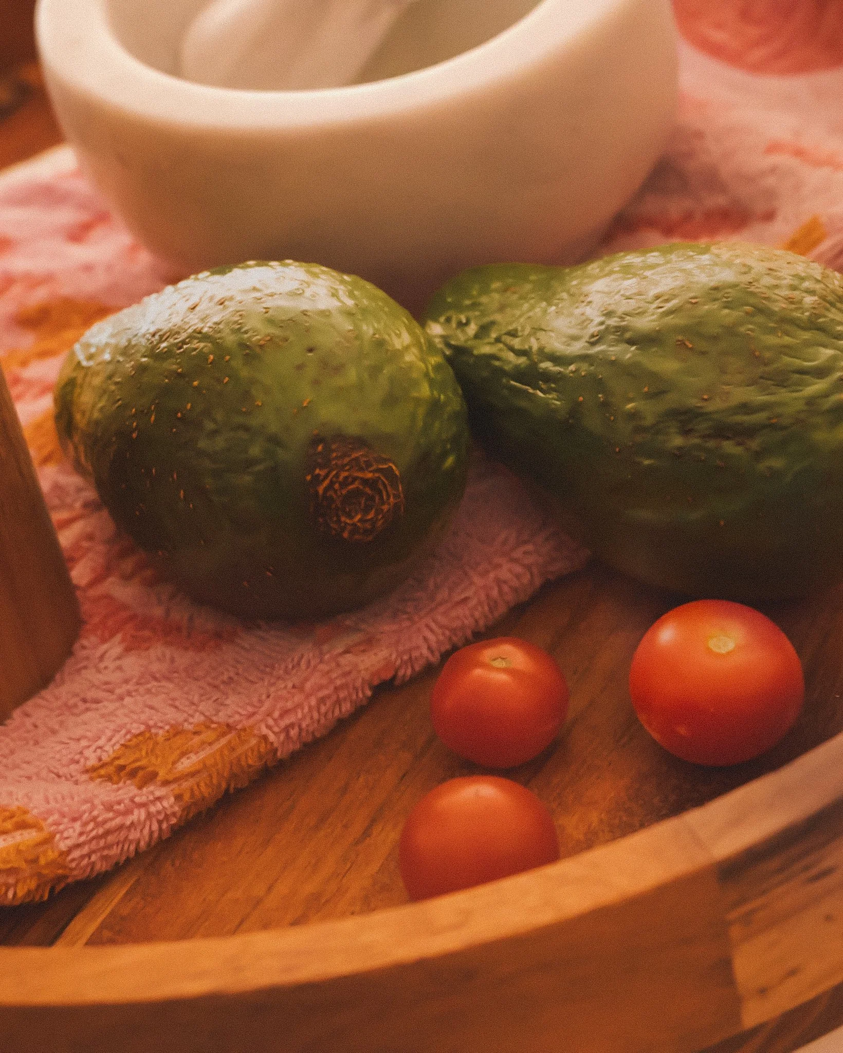 Two avocados, three cherry tomatoes, a white bowl, and a pink cloth on a wooden tray.