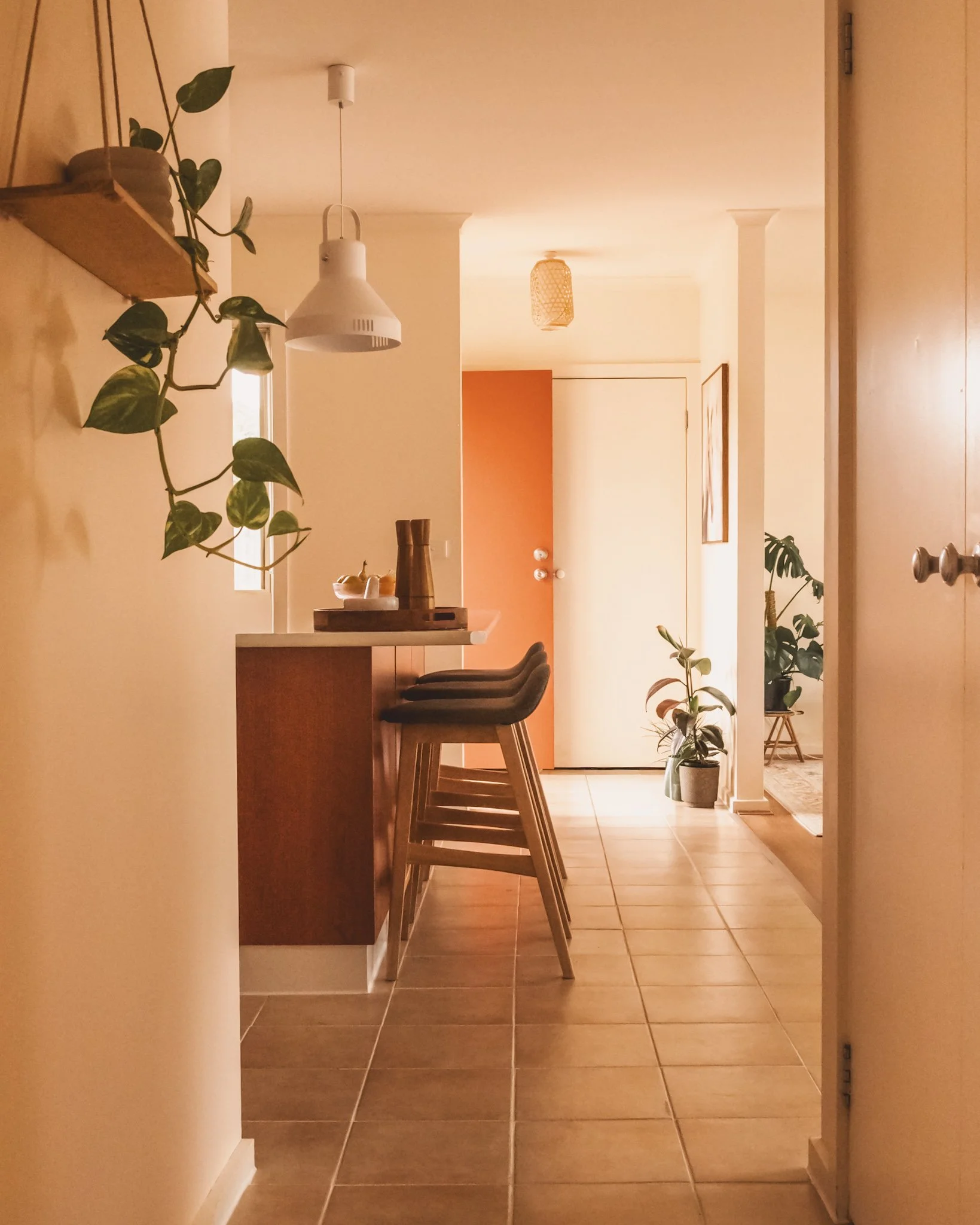 A cozy, well-lit hallway with a kitchen counter on the left side and several bar stools tucked in. There are potted plants placed along the hallway, with a door at the end. The decor features warm tones, and the space appears welcoming and modern.