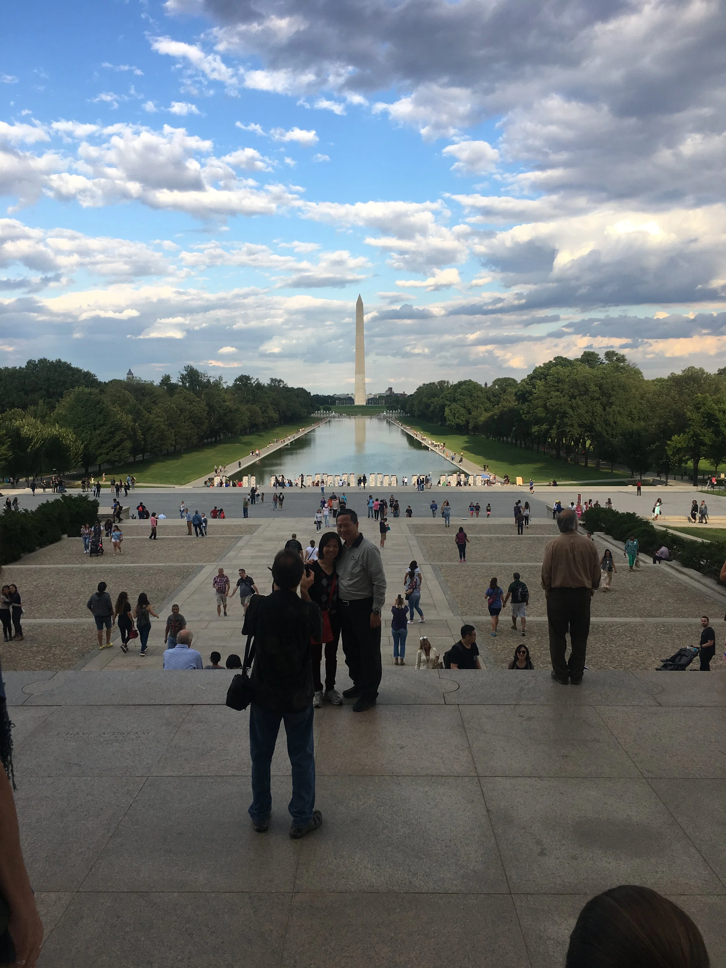 The photo shows the Washington Monument in Washington, D.C., with a reflecting pool in front, trees on either side, and people walking or standing on the steps leading down to the pool on a partly cloudy day.