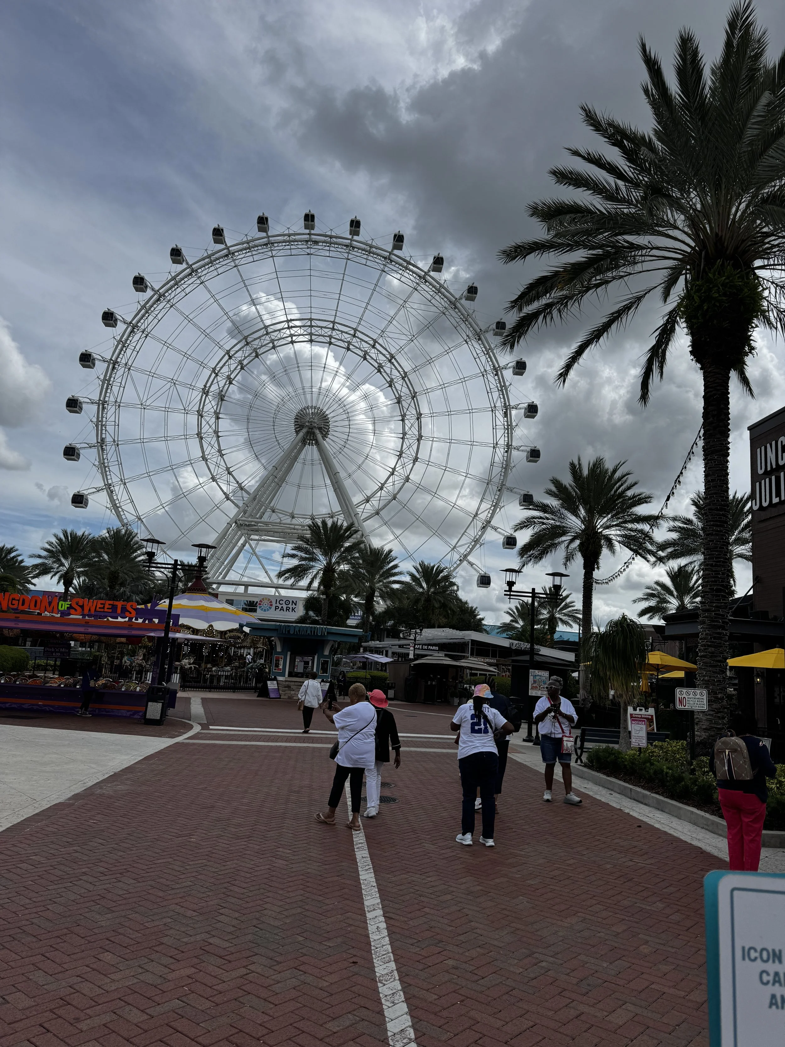 People walking in front of the Eye at ICON Park win Orlando, Florida with a large Ferris wheel, palm trees, and cloudy sky in the background.