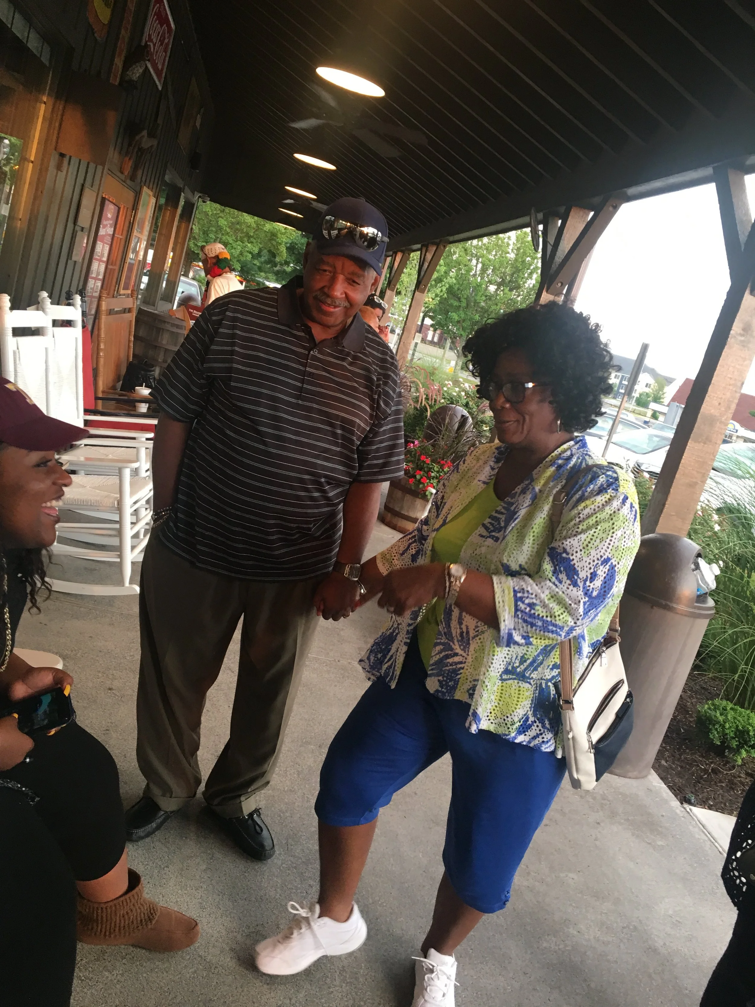 Two women and one man standing on a porch, engaging in conversation and hand-holding, with one woman sitting nearby.