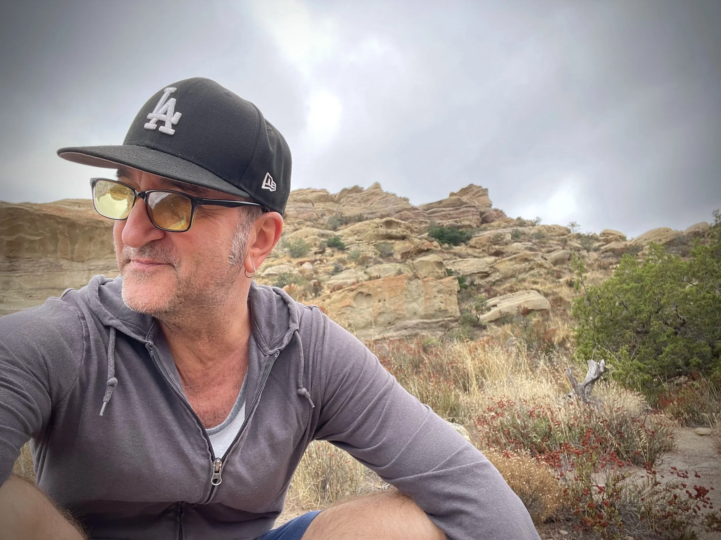 A man with sunglasses, a black cap, and a gray zip-up hoodie sitting in a desert landscape with rocky formations, shrubs, and a cloudy sky.