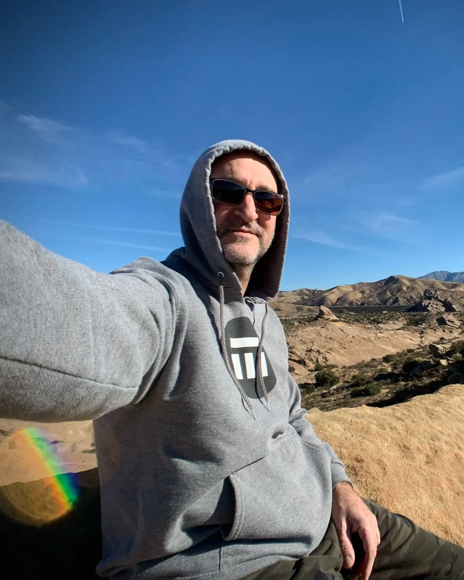 A man wearing sunglasses and a gray hoodie taking a selfie outdoors with a mountainous rocky landscape and a blue sky in the background.