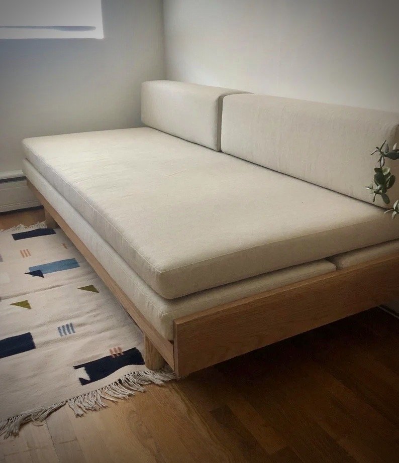 A minimalist bedroom with a beige bed, a light-colored wooden frame, and a patterned rug on hardwood flooring.