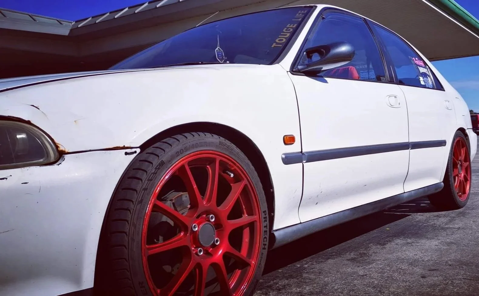 A white car with red rims, showing front left side, parked under a bridge or structure, with some visible damage and rust near the fender.