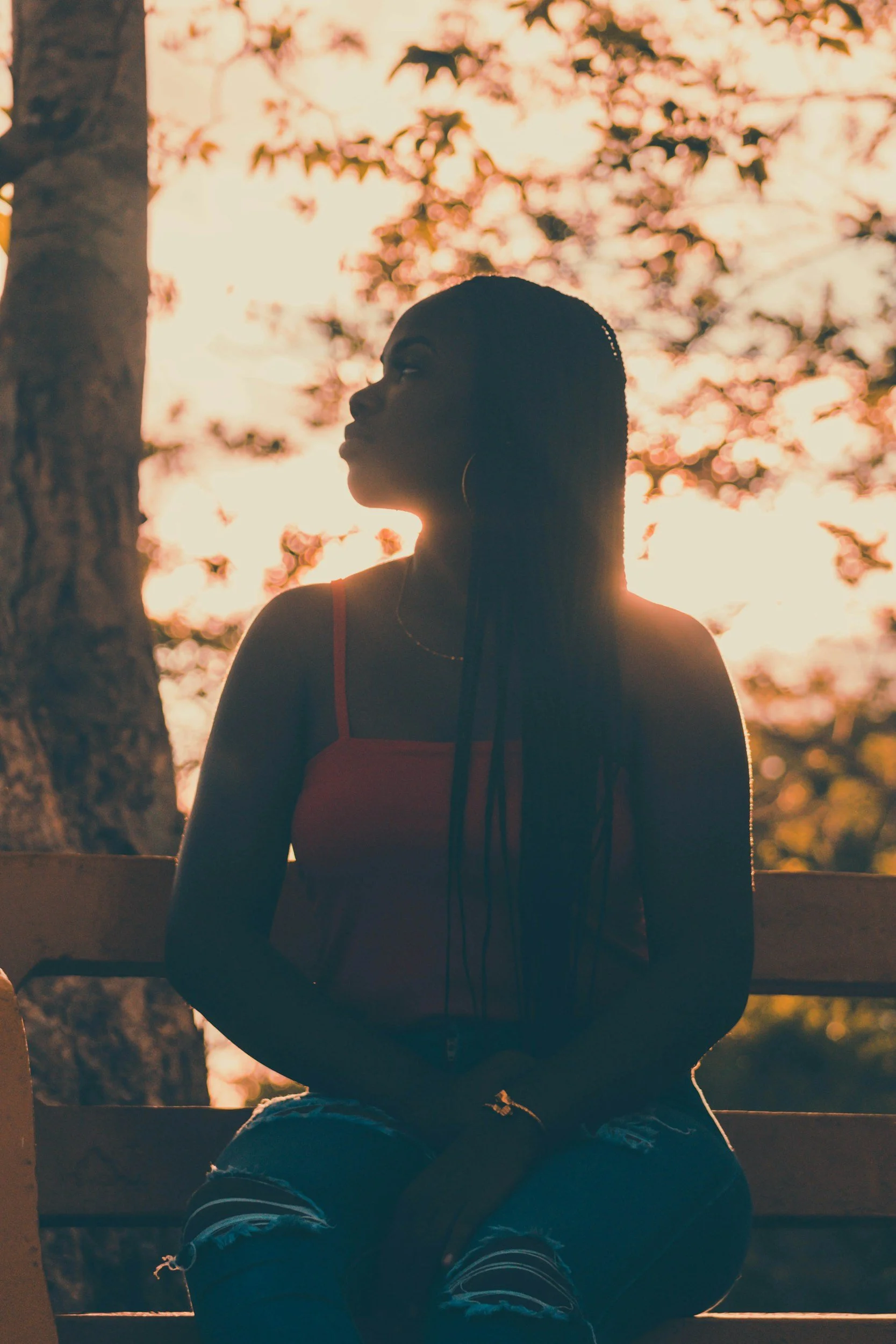 Silhouette of a woman sitting on a park bench during sunset, with her face turned to the side against a background of trees and a warm sky.