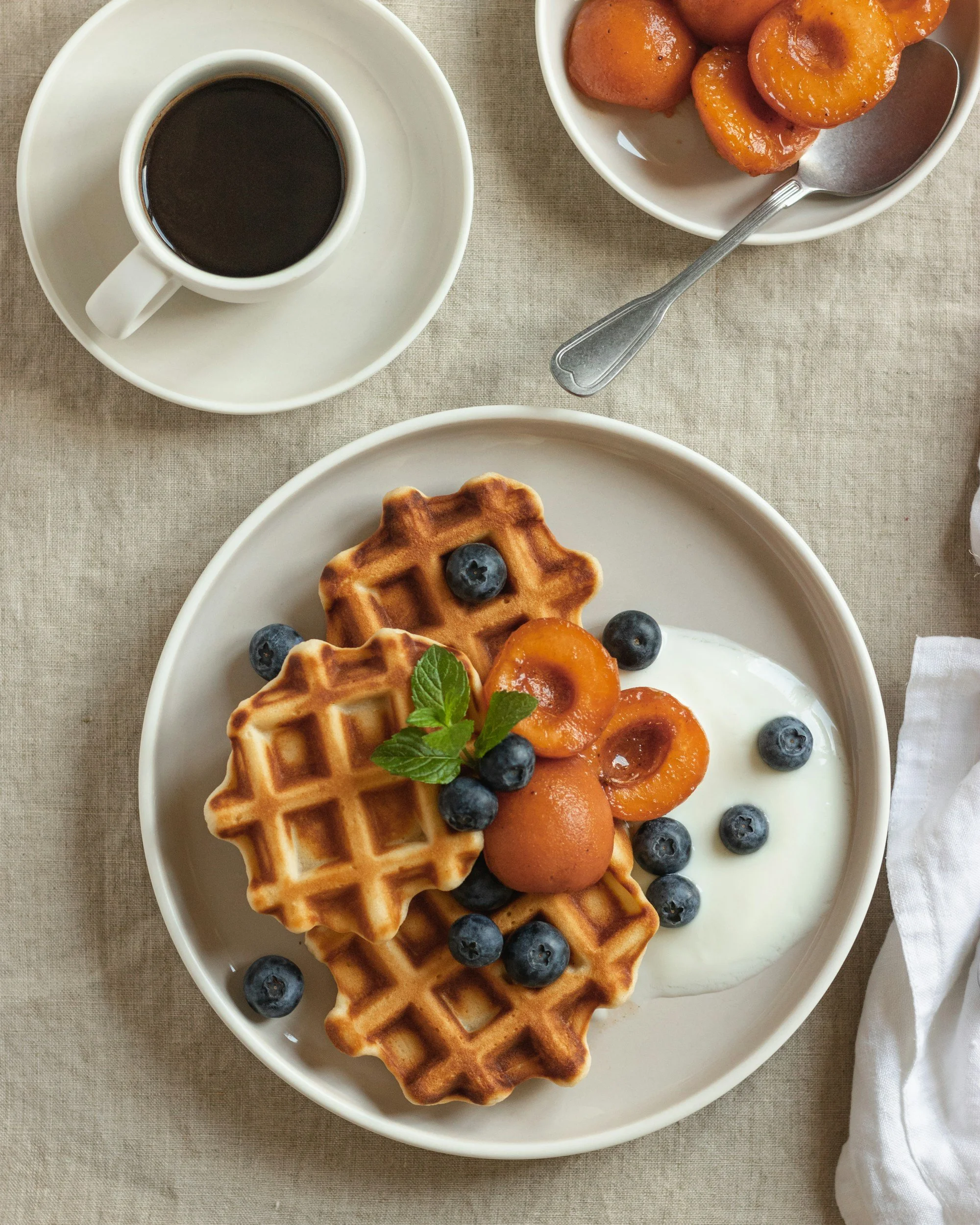 Plate of waffles topped with blueberries, apricots, a sprig of mint, and a dollop of cream, with a cup of coffee and a bowl of apricots on the side.