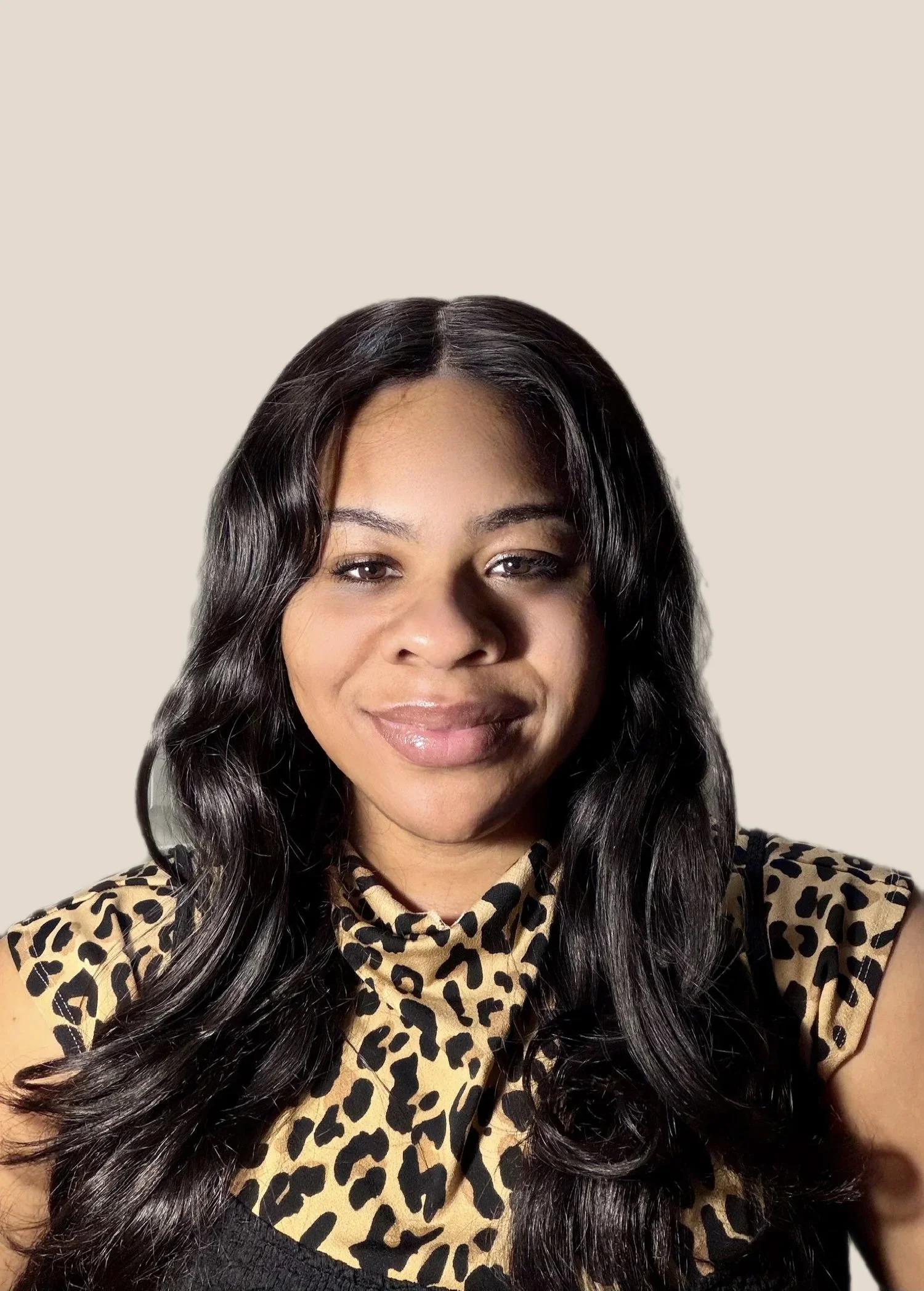 A woman with long black wavy hair smiling, wearing a leopard print top, in front of a plain wall with a door visible in the background.