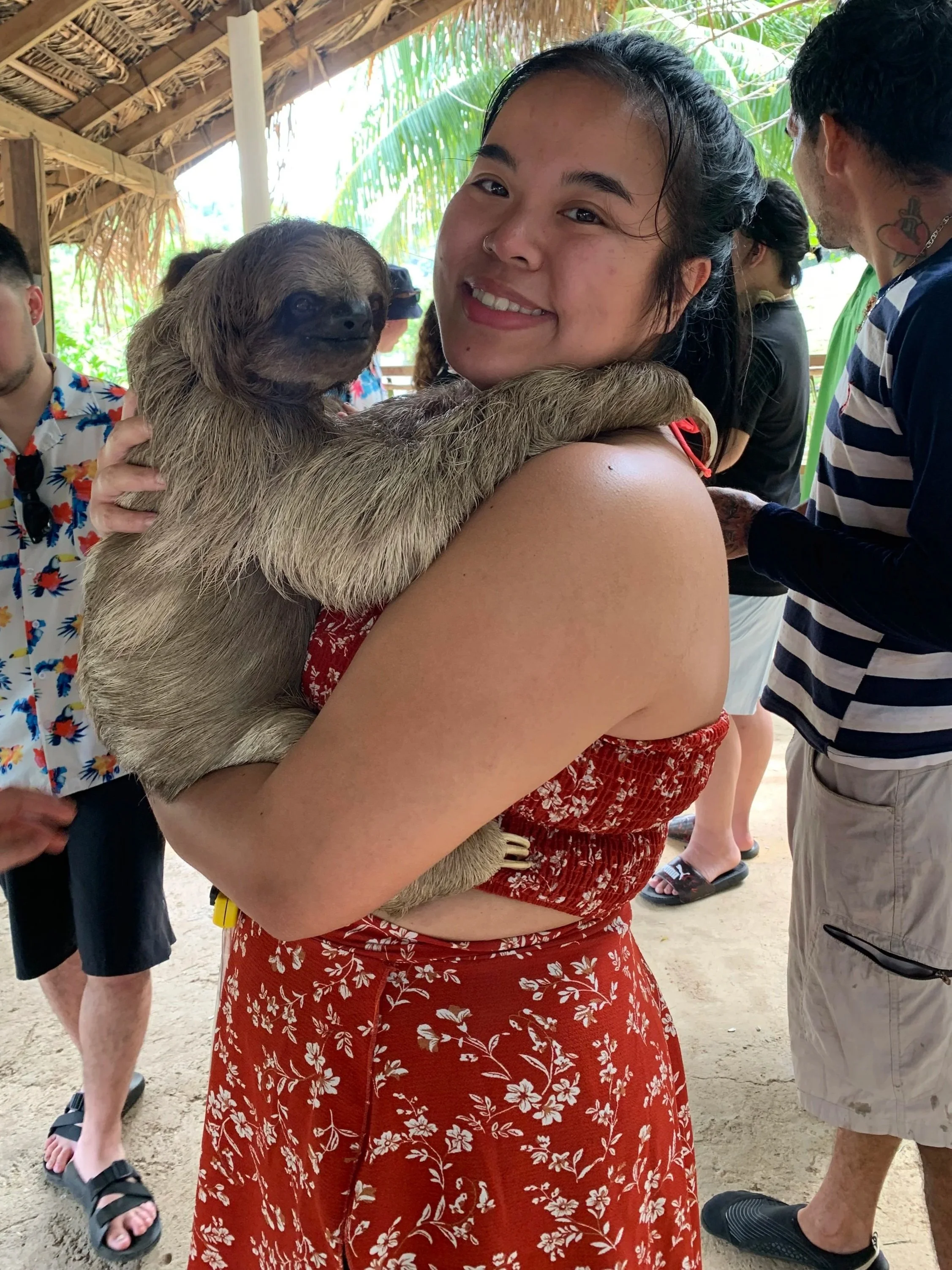 A young woman in a red floral dress is holding a sloth on her shoulders, smiling at the camera, in a tropical setting with people in the background.
