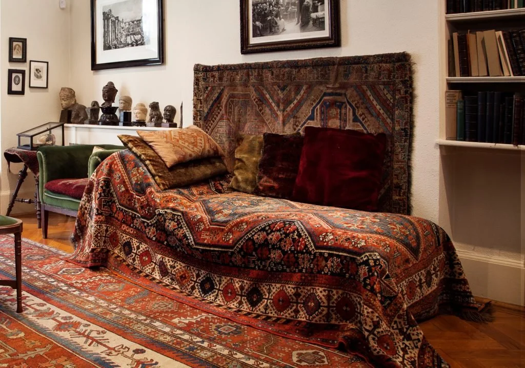 A living room corner with a red and orange patterned Ottoman-style sofa, decorated with a few velvet cushions, a small green velvet armchair, a bookshelf filled with books, and framed pictures and artwork on the walls. There are also bust sculptures on a white console against the wall.