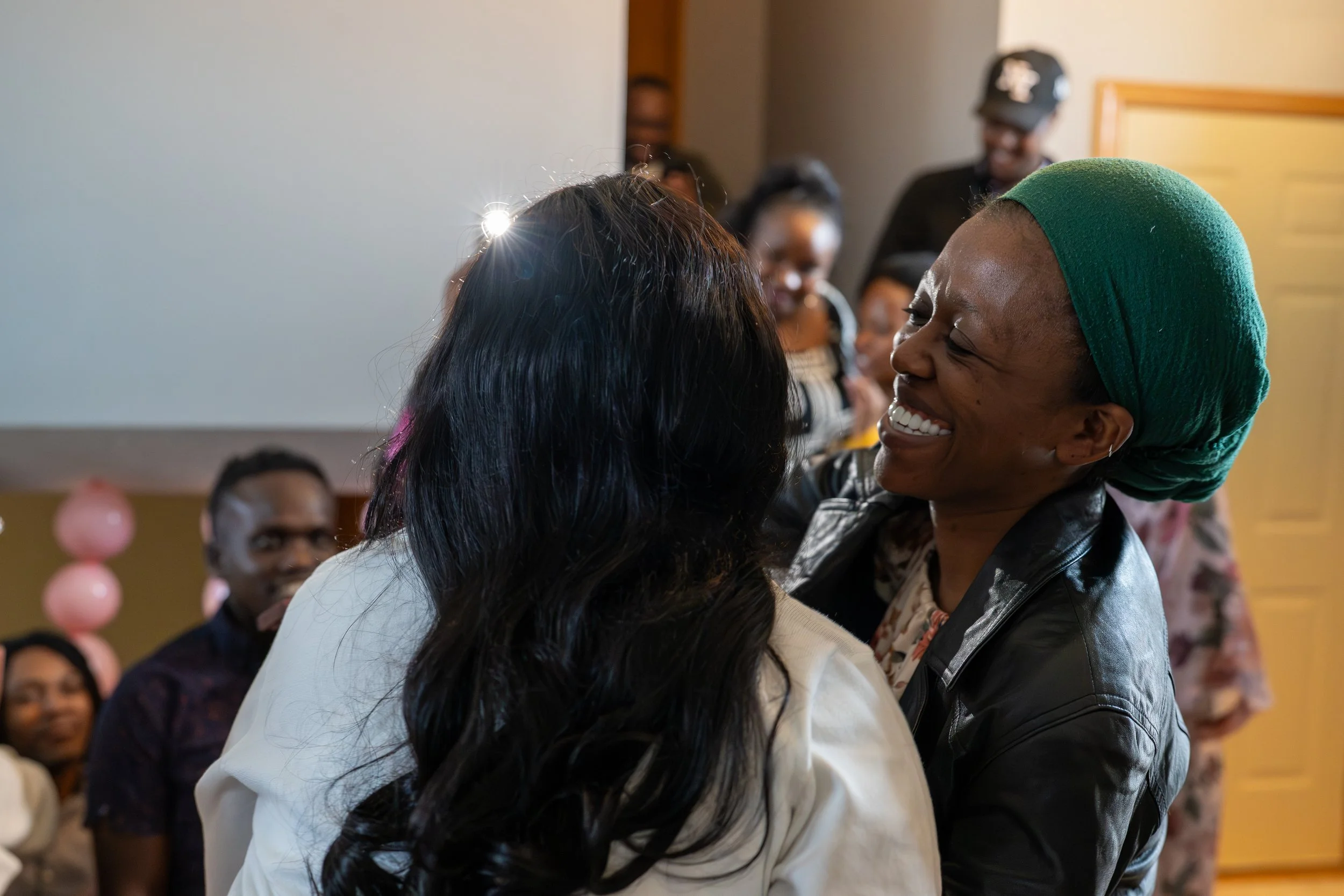 Two women share a joyful hug at a celebration with people in the background, including a man wearing glasses and women smiling.
