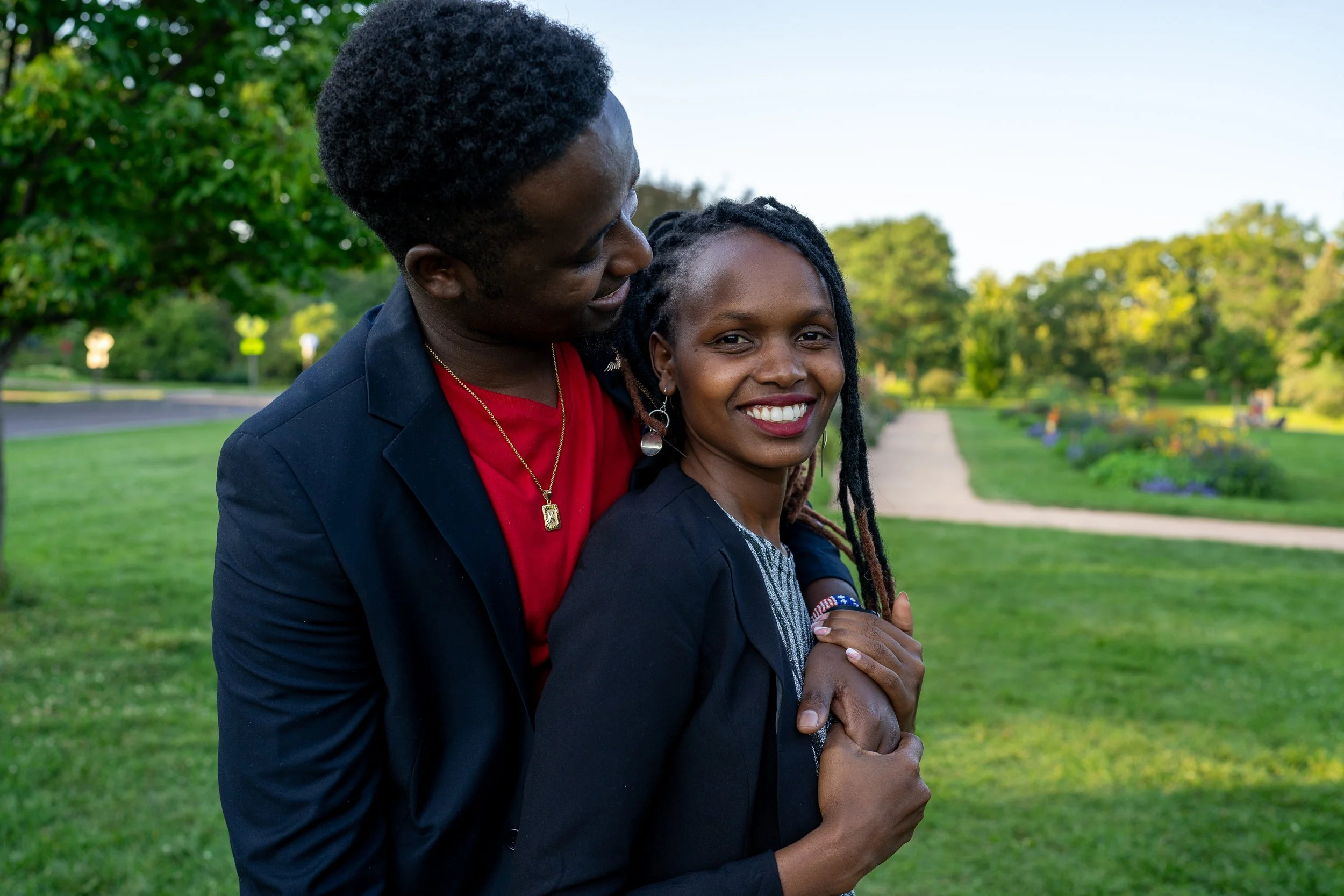 A happy Black woman with dreadlocks and a black blazer smiling at the camera, standing outdoors in a park, while a Black man with short curly hair and a navy blazer stands close behind her, looking at her affectionately during the daytime.