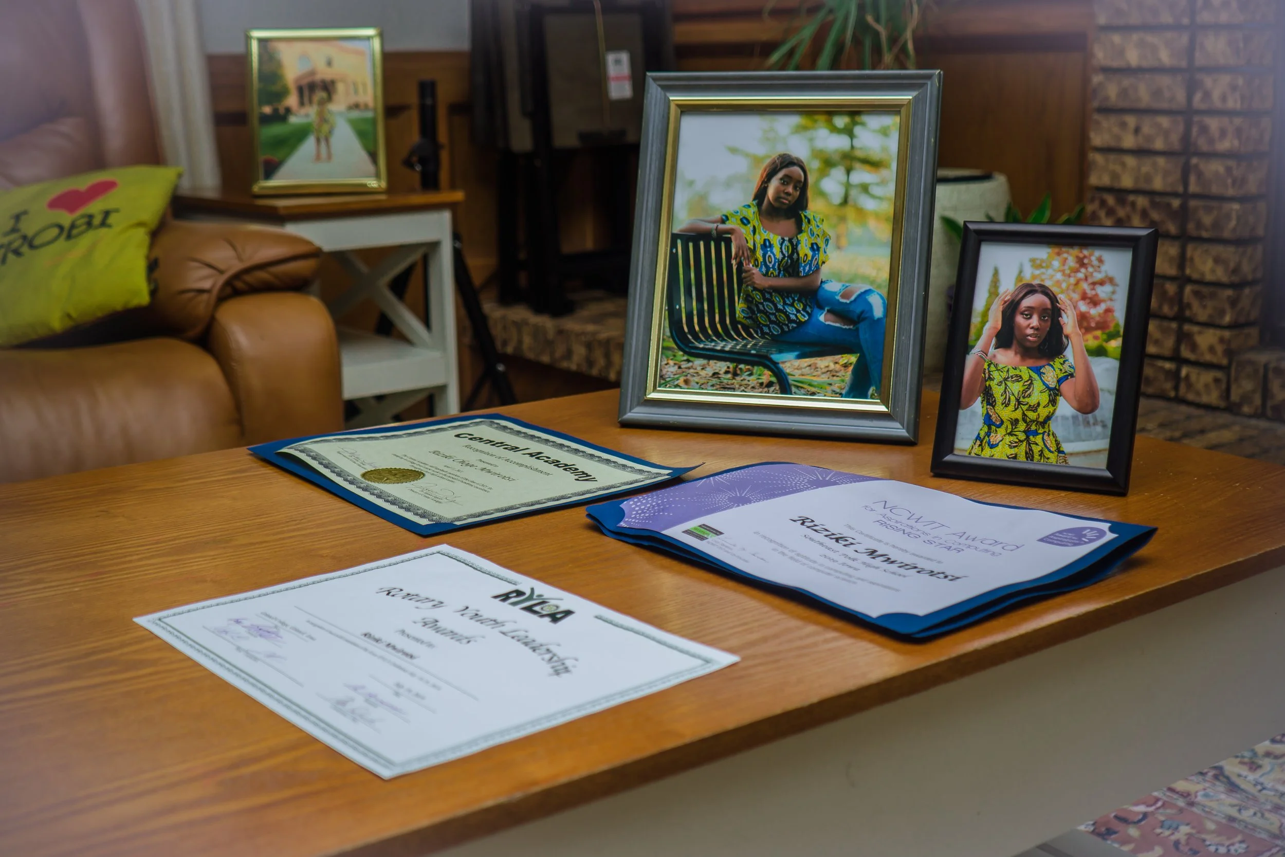 A wooden table featuring diplomas and framed photographs of a young woman, with a couch and decorative pillows in the background.