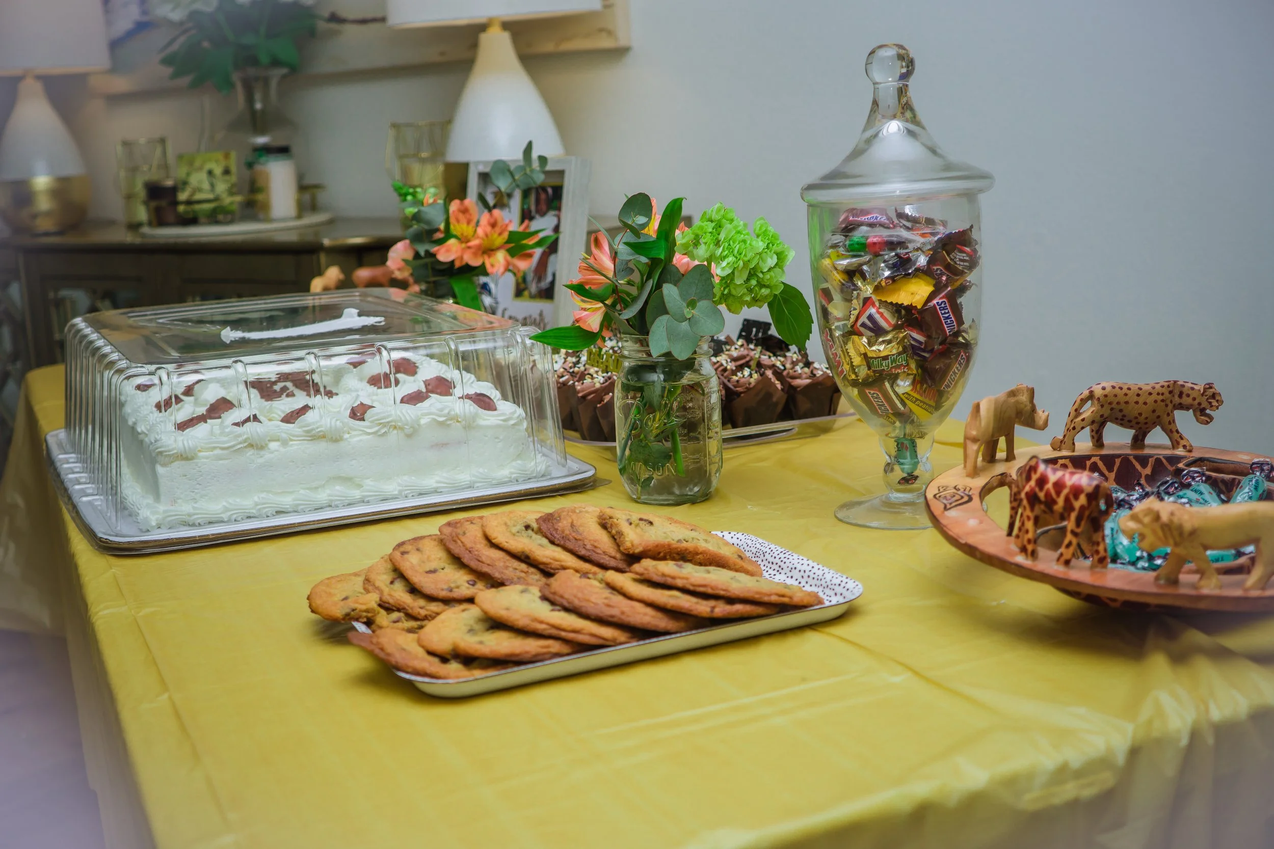 Table with a yellow tablecloth holding a decorated cake under a clear plastic cover, a tray of cookies, a small vase of pink and green flowers, a large jar of assorted candies, and a decorative tray with toy animal figurines.