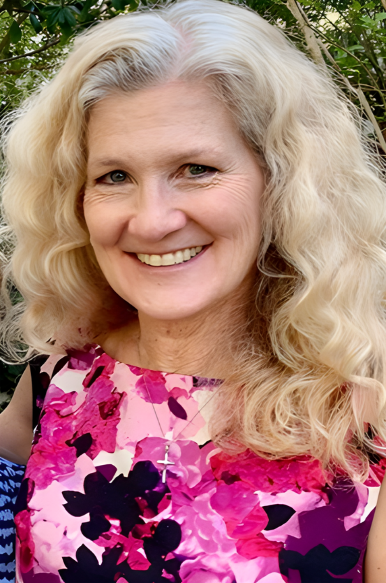 A woman with curly blonde hair smiling outdoors, wearing a pink and purple floral dress with a black background.
