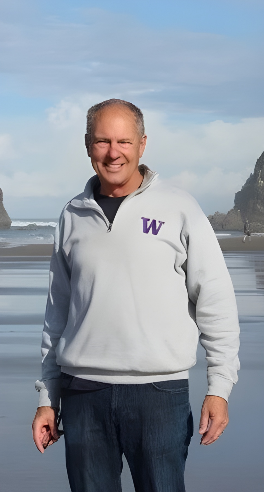 A smiling elderly man standing on a beach with rocks and ocean waves in the background, wearing a light gray sweatshirt with a purple "W" on the chest and dark pants.