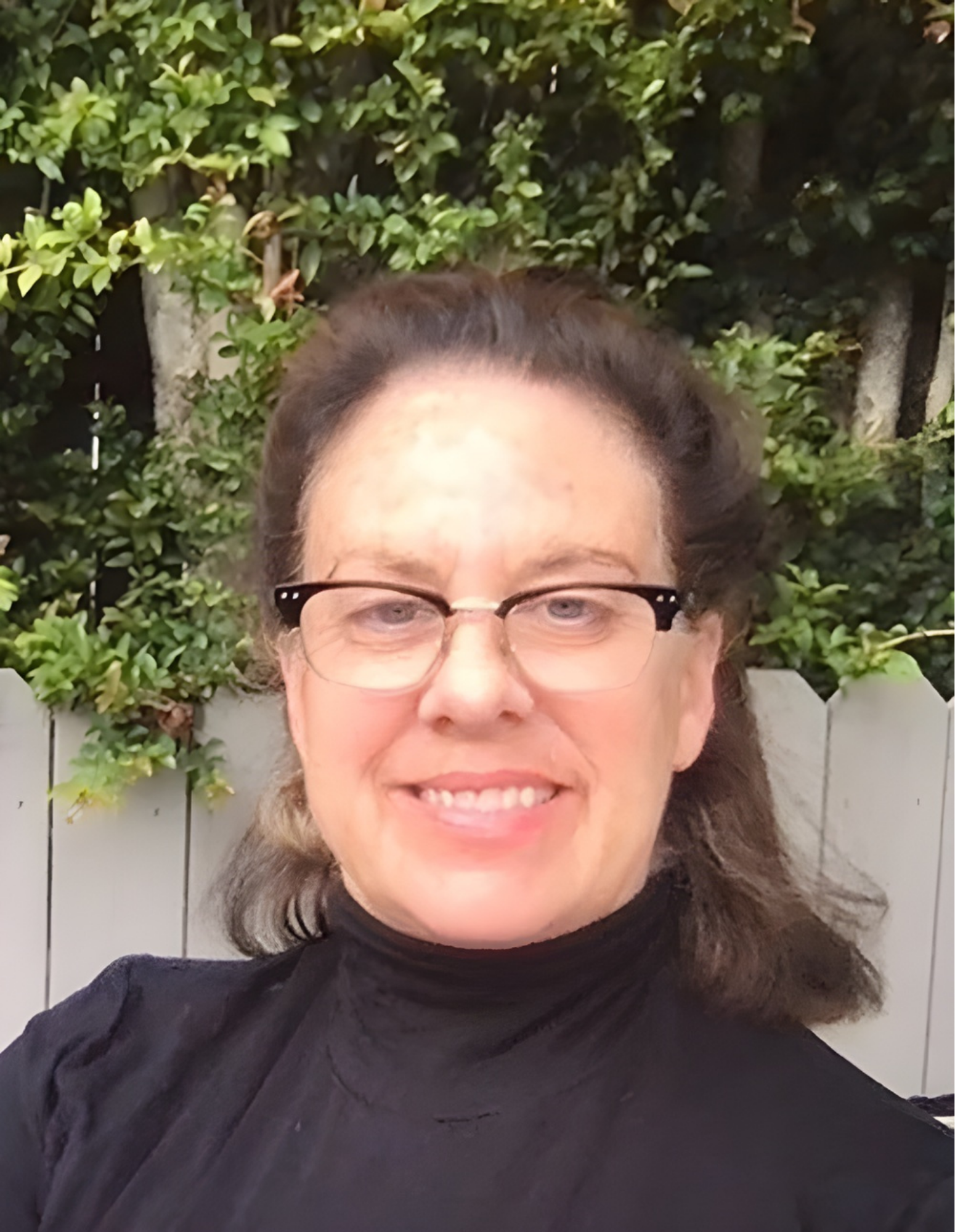A woman with glasses and shoulder-length hair, wearing a black turtleneck, standing outdoors in front of greenery and a wooden fence.