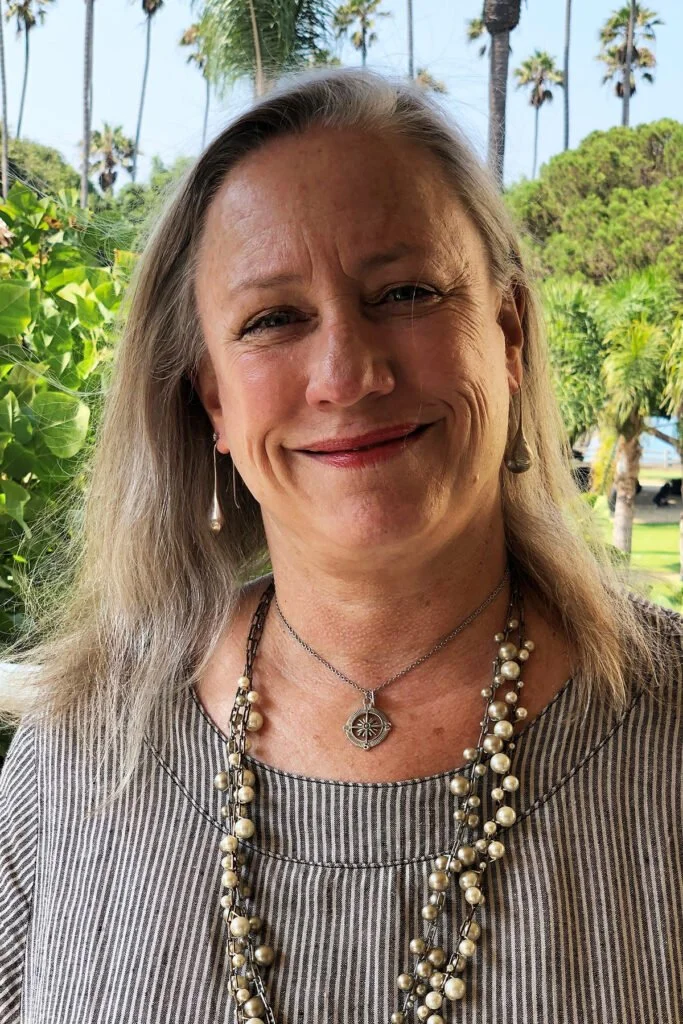 A smiling woman with long blonde hair, wearing pearl necklaces and earrings, standing outdoors with palm trees and greenery in the background.