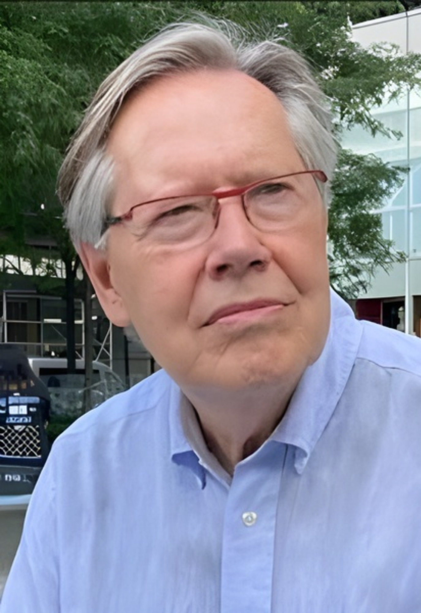 Older man with gray hair and glasses, wearing a light blue shirt, standing outdoors near a modern glass building and trees.