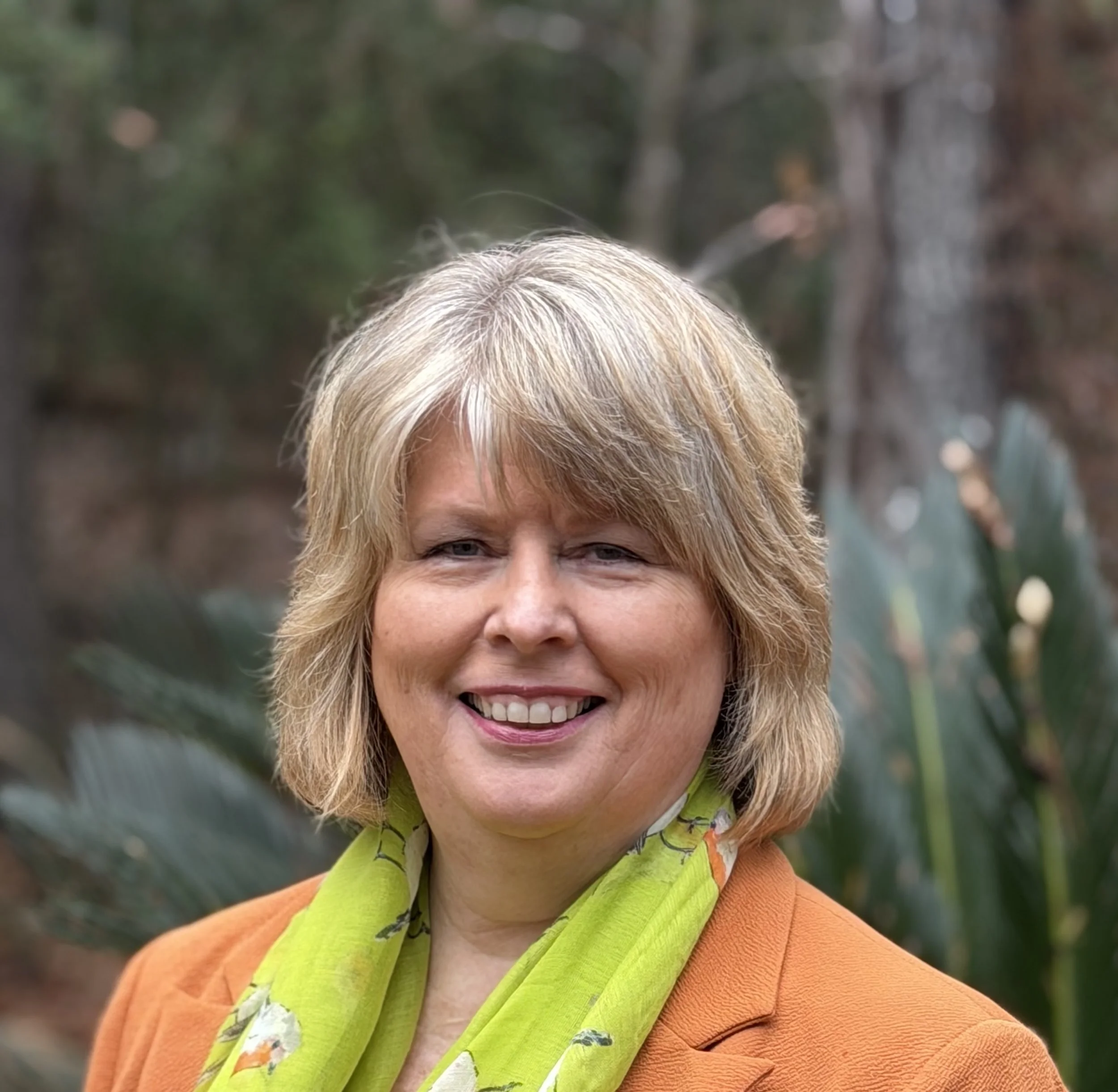 Smiling woman with short light brown hair, wearing large round earrings and a pink blazer, sitting in front of an abstract colorful painting.