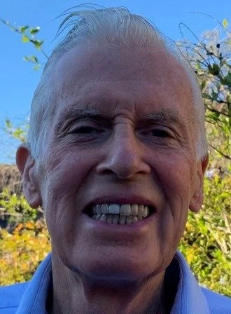 Close-up of an elderly man smiling outdoors with a blue sky and green foliage in the background.