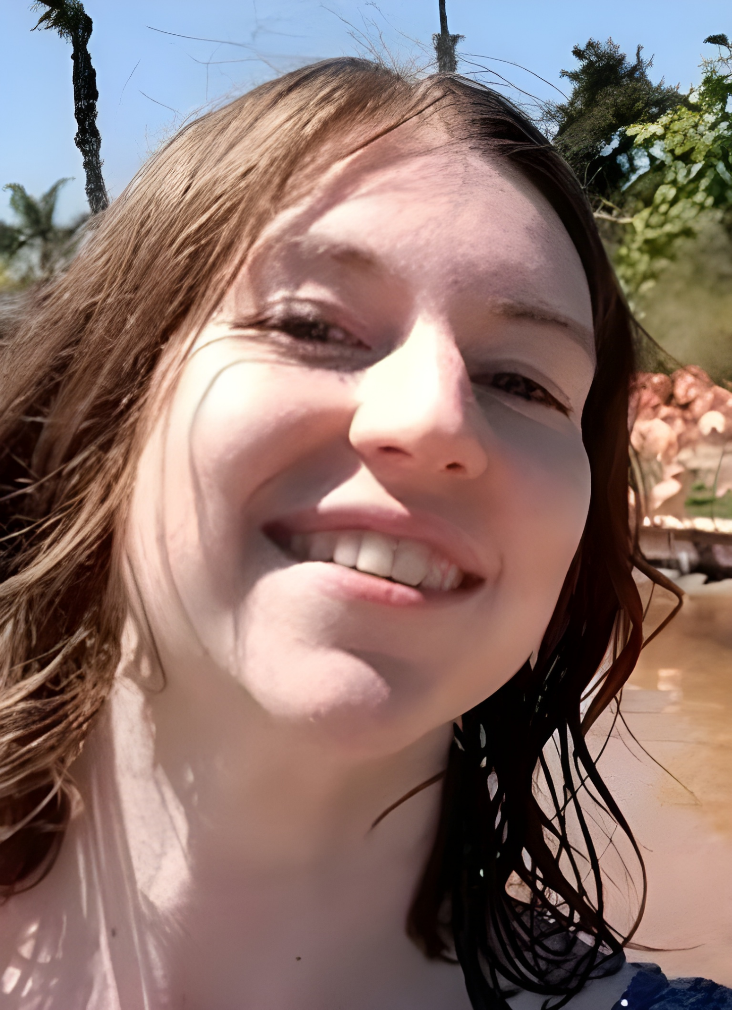 Close-up of a smiling woman with long brown hair outdoors on a sunny day.