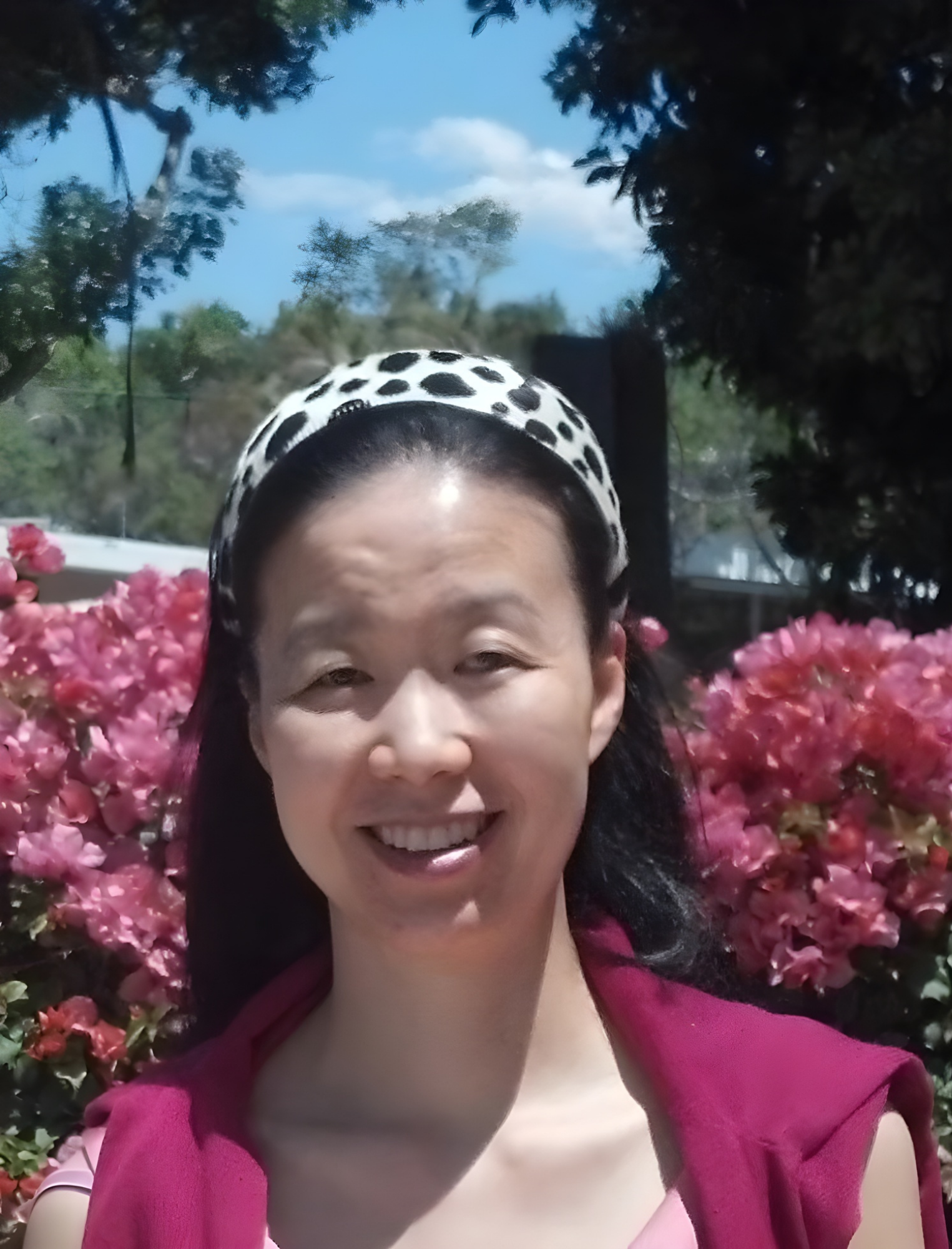 A woman with long dark hair wearing a white and black polka dot headband, smiling, standing outdoors in front of pink flowering bushes, with blue sky and trees in the background.