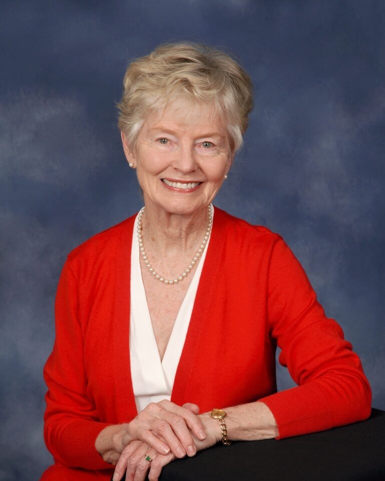 An elderly woman with short, light-colored hair, smiling, wearing a red cardigan over a white top, a pearl necklace, and a gold bracelet, sitting with her hands clasped on a table in front of a blue background.