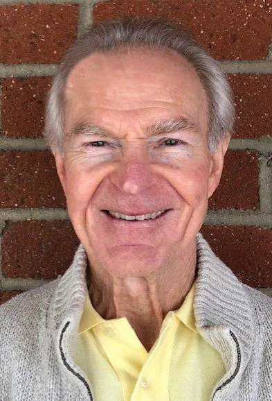 An elderly man with gray hair, smiling, standing in front of a brick wall, wearing a light-colored sweater over a yellow collared shirt.
