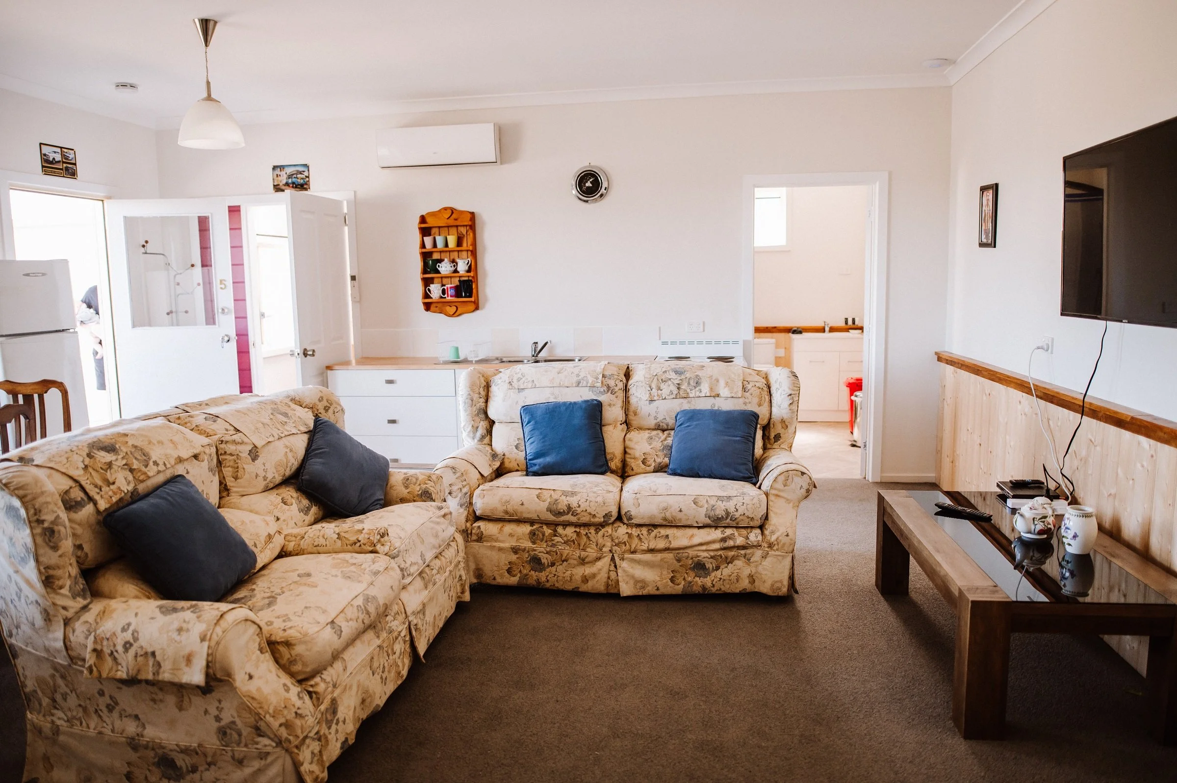 Living room with floral sofas, blue pillows, wall-mounted TV, wooden coffee table, open kitchen in the background, and a doorway leading to another room.