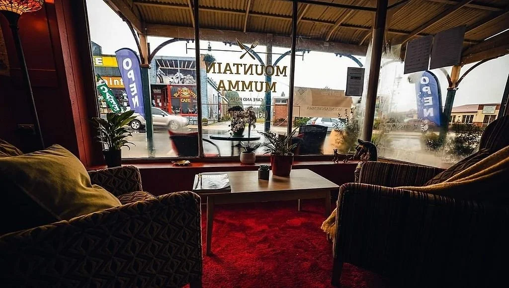 Cozy indoor cafe viewed through large front window, with plants on the windowsill, two striped armchairs and a table inside, and outdoor signs for 'Nero' and others visible outside.