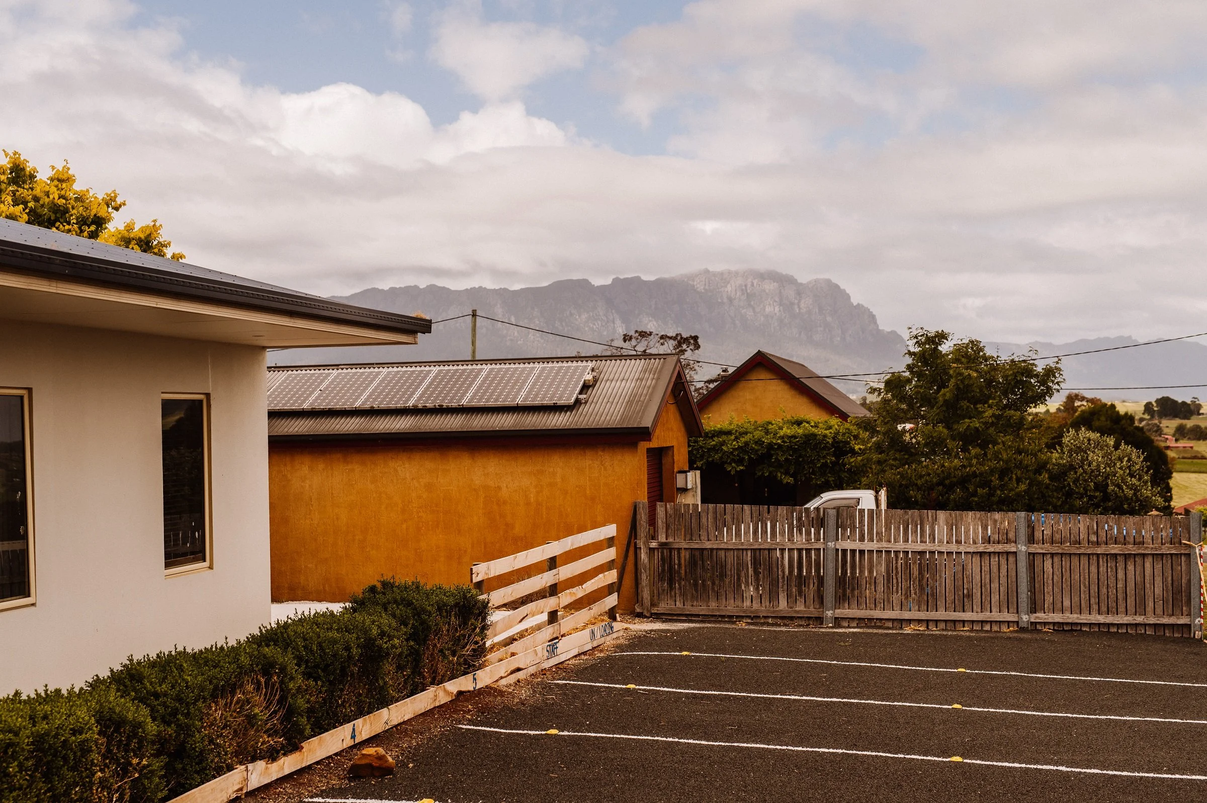 A parking lot with white lines, a wooden fence, green bushes, and behind it a tan shed, a white house, trees, and mountains in the background under a partly cloudy sky.