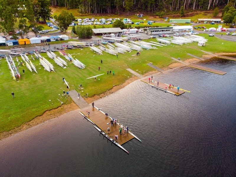 Aerial view of a boat marina with sailboats on grassy land, people walking near the shore, and a dock extending into the water at a boat launch area.