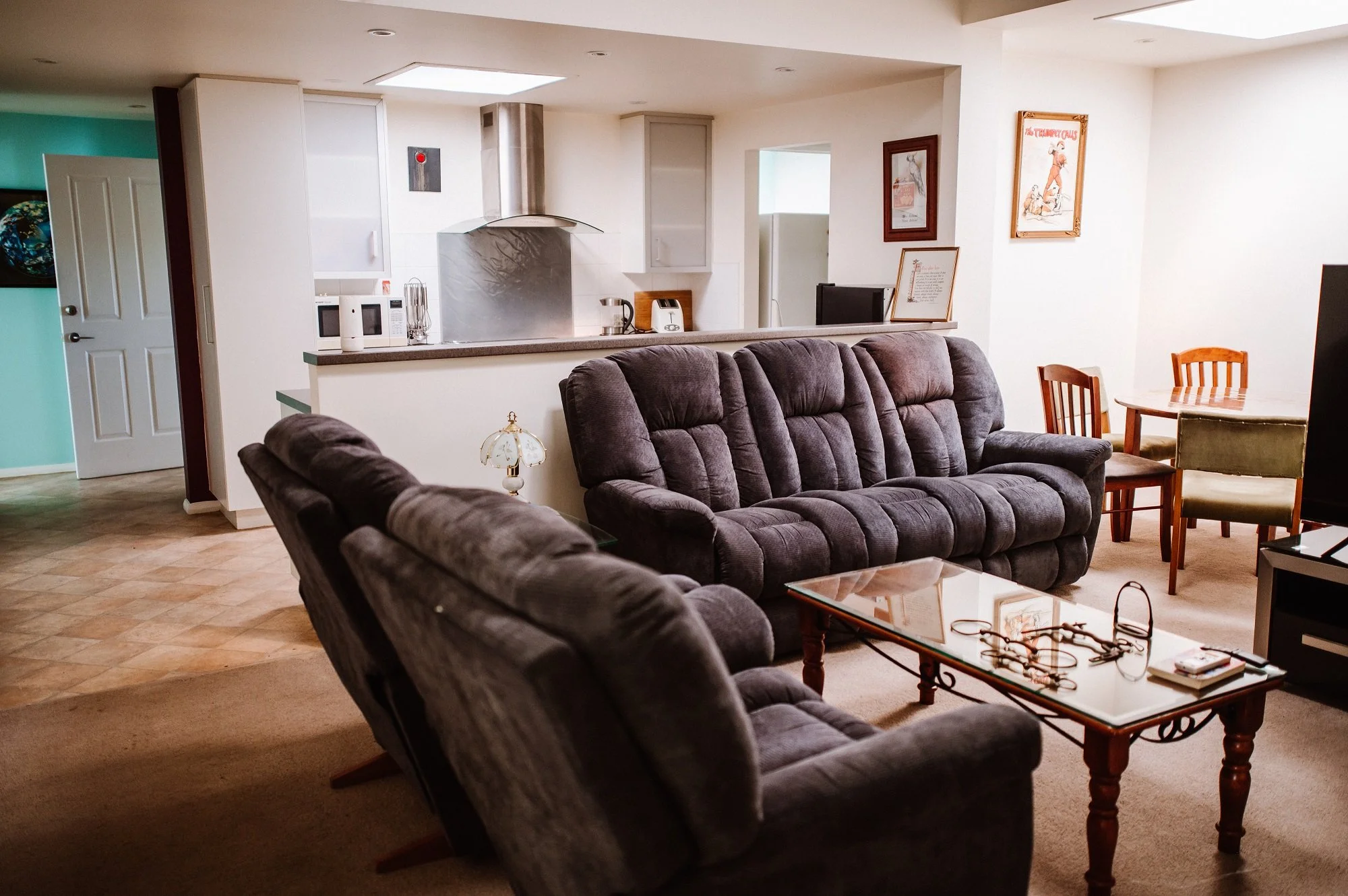 Living room with dark gray upholstered sofas, a glass-topped wooden coffee table, and a partial view of the kitchen in the background with white cabinets, a stainless steel hood, and appliances. There is a dining table with chairs on the right.