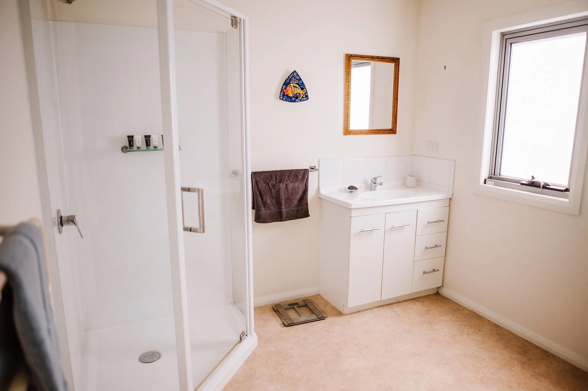 Bathroom with a glass shower enclosure, a white vanity with sink, a brown towel hanging on the wall, a small mirror, and a window letting in natural light.