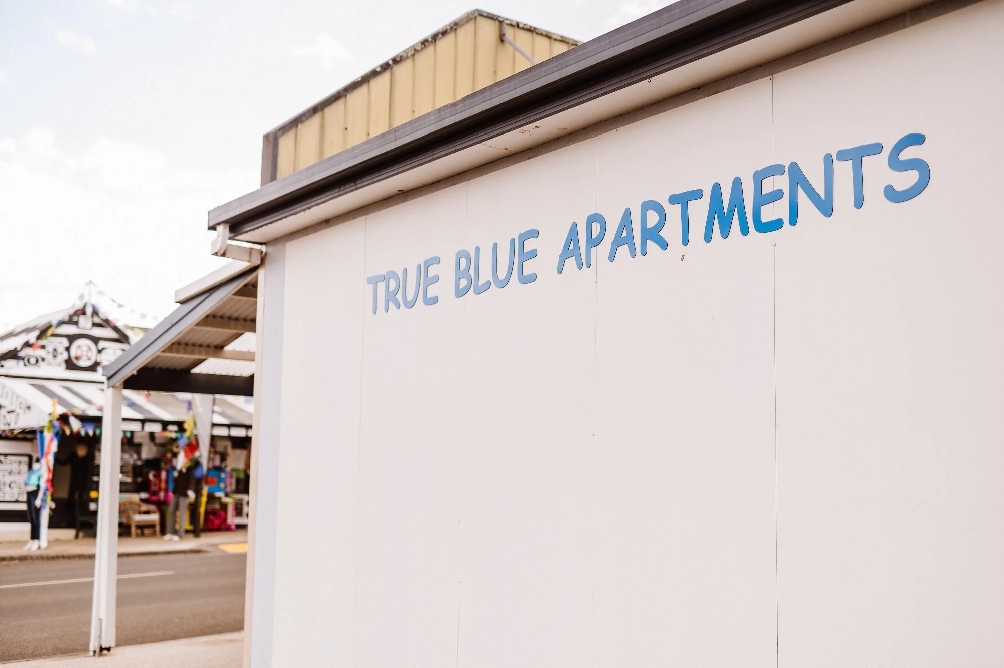 Sign on a white building reading 'True Blue Apartments,' with an outdoor market in the background featuring tents and people.