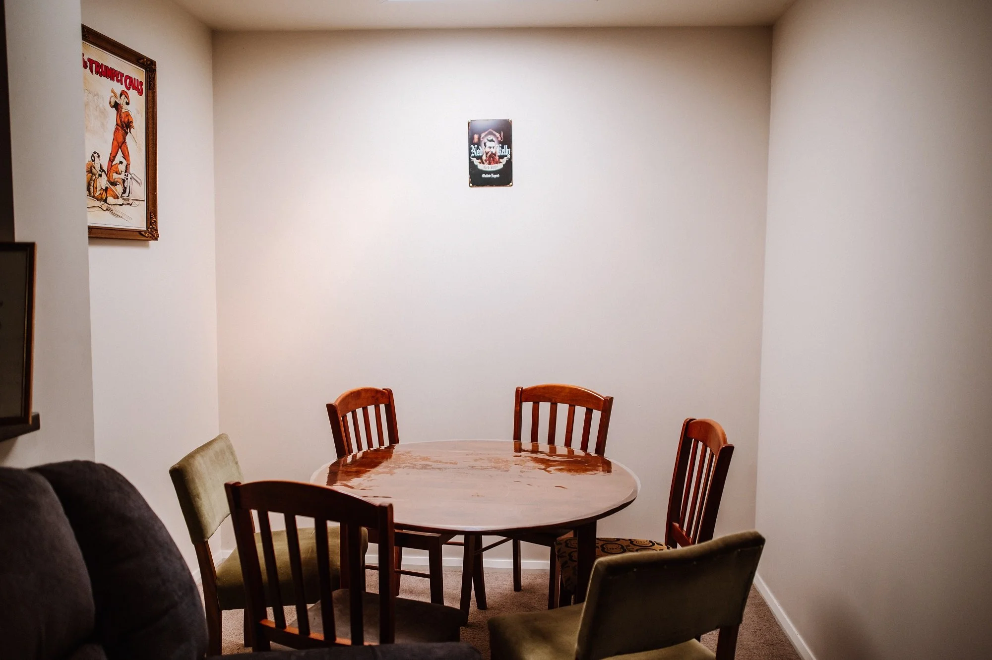 Empty dining room with a round wooden table surrounded by five chairs, two of which are upholstered in green, and three in wood finish. The room has plain white walls, two wall posters, with one on the left depicting a rodeo scene and one on the center back showing a skull face. There is a dark sofa partially visible on the left side. The floor is carpeted in beige.